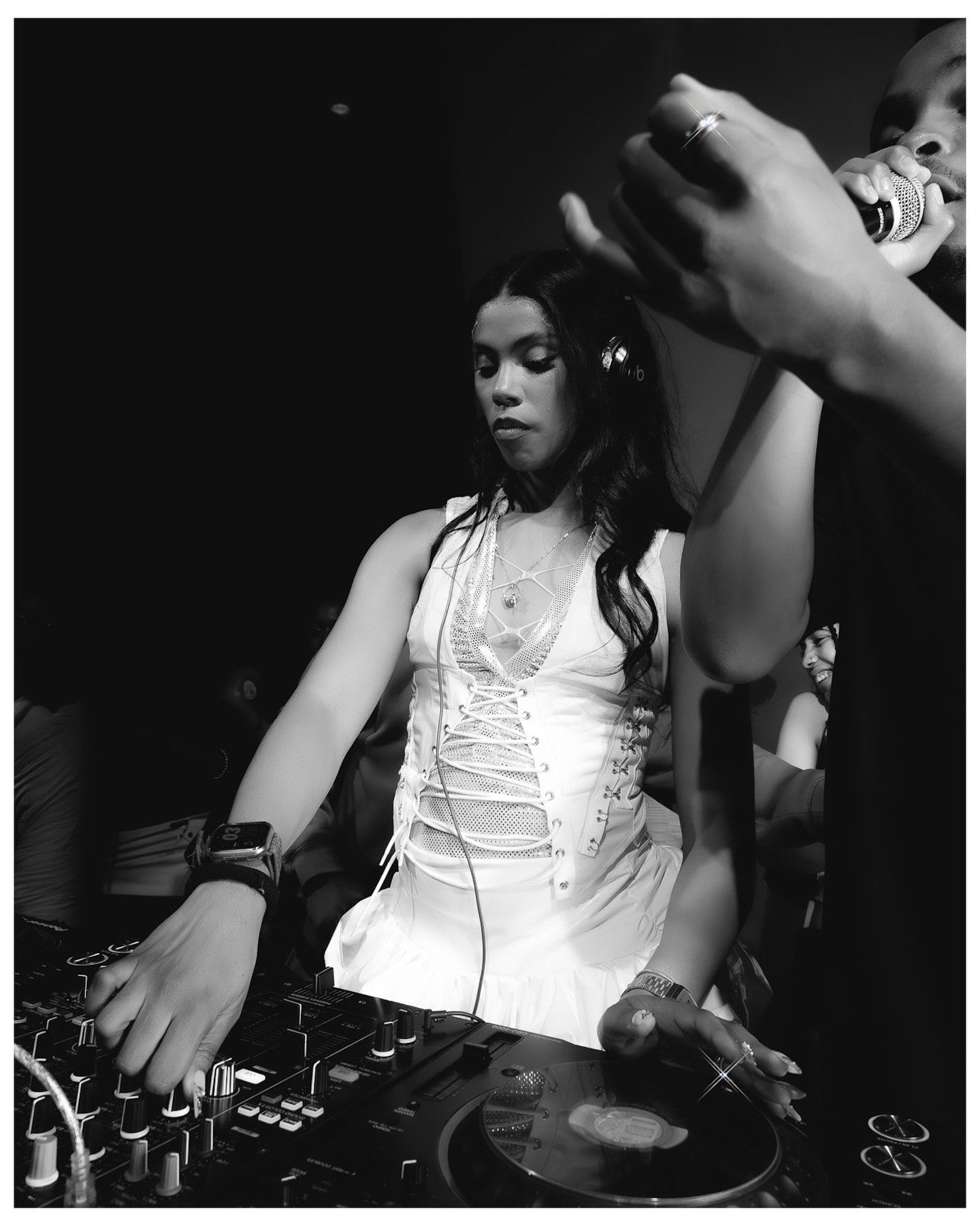 Female DJ in white dress mixing at turntables in black and white club photo.