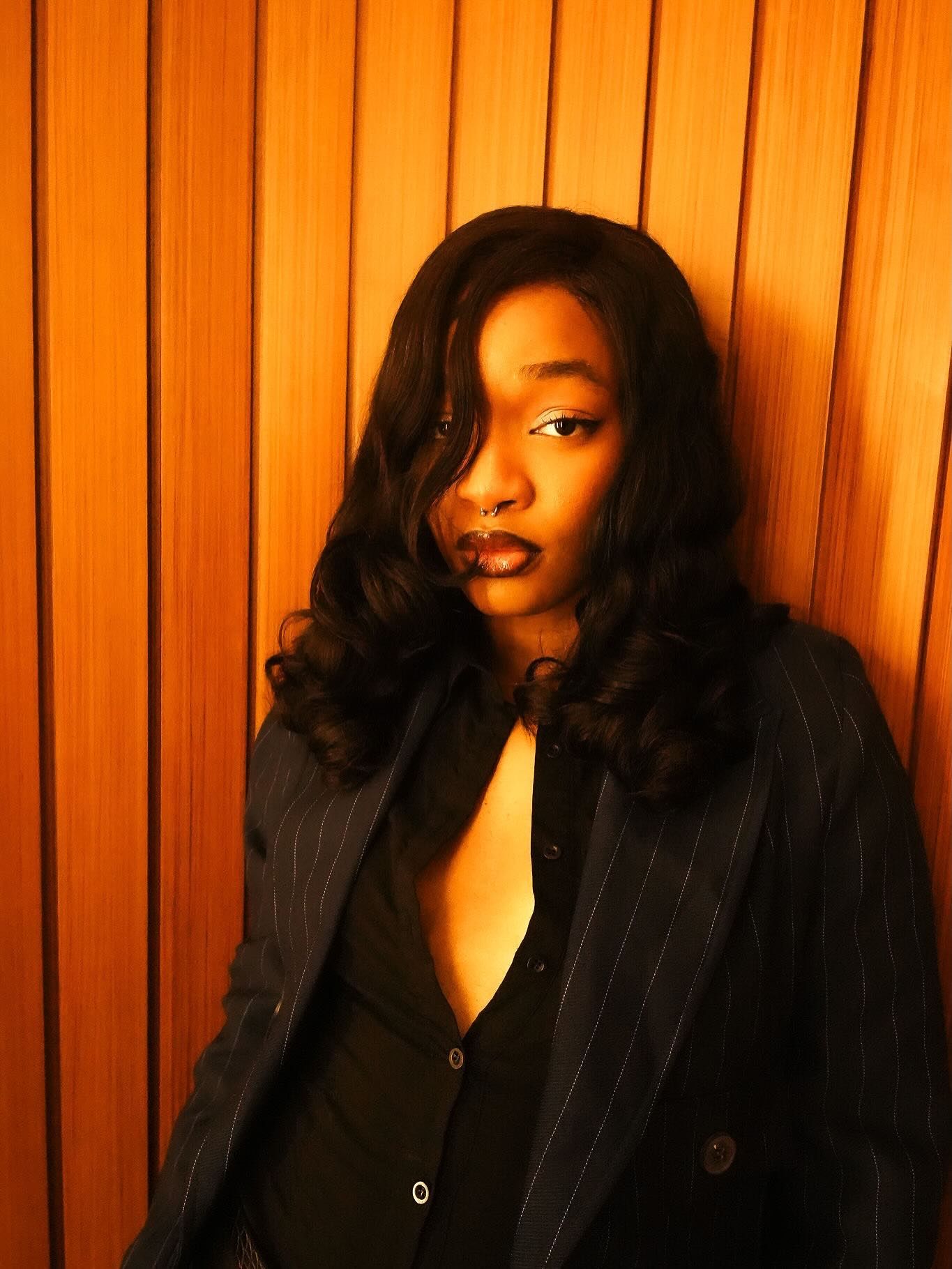 Young Black woman in pinstripe blazer poses against warm wood-paneled wall with curly hair and nose ring.