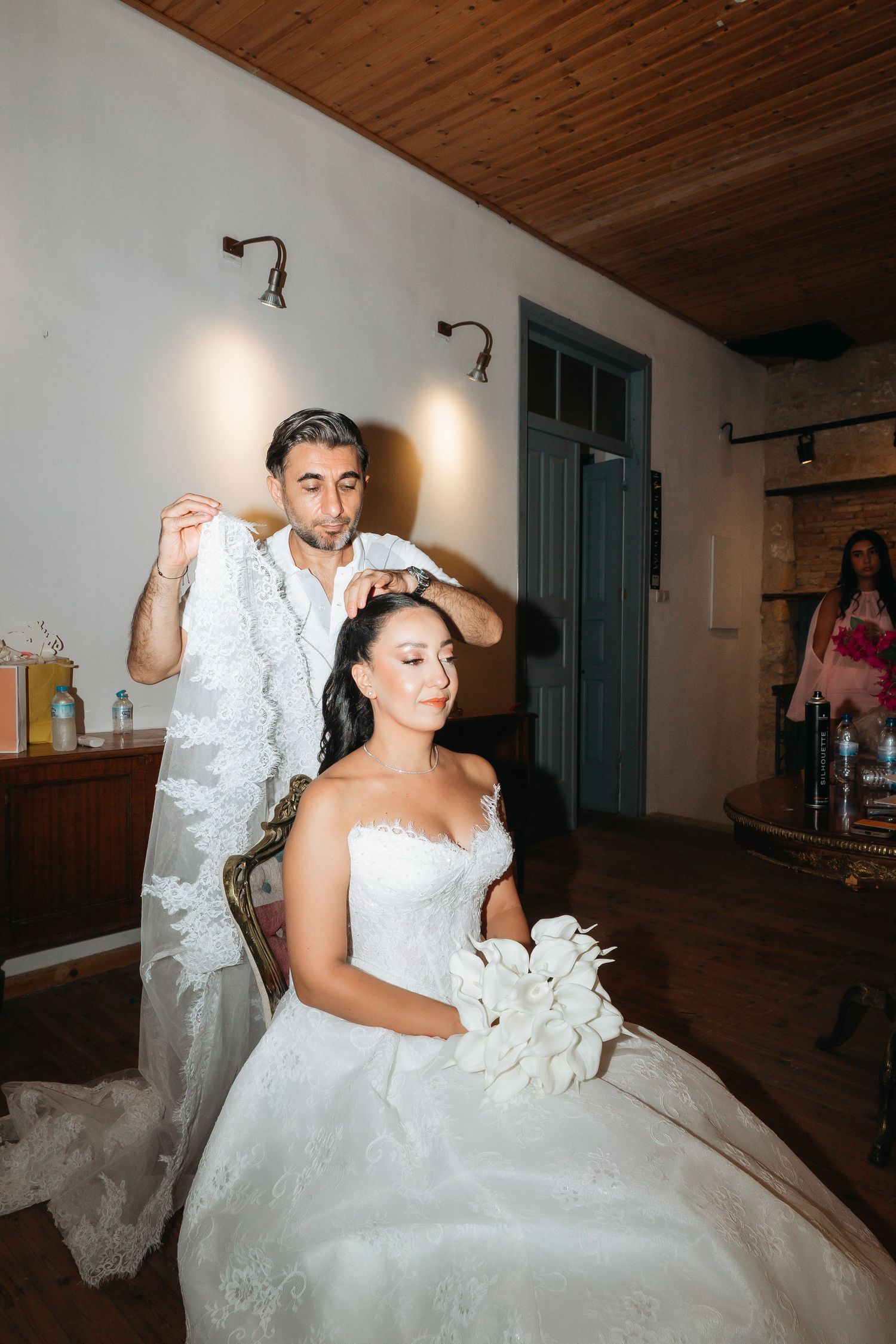 A hairstylist carefully places a lace veil on a seated bride in a white gown holding a bouquet before her wedding.