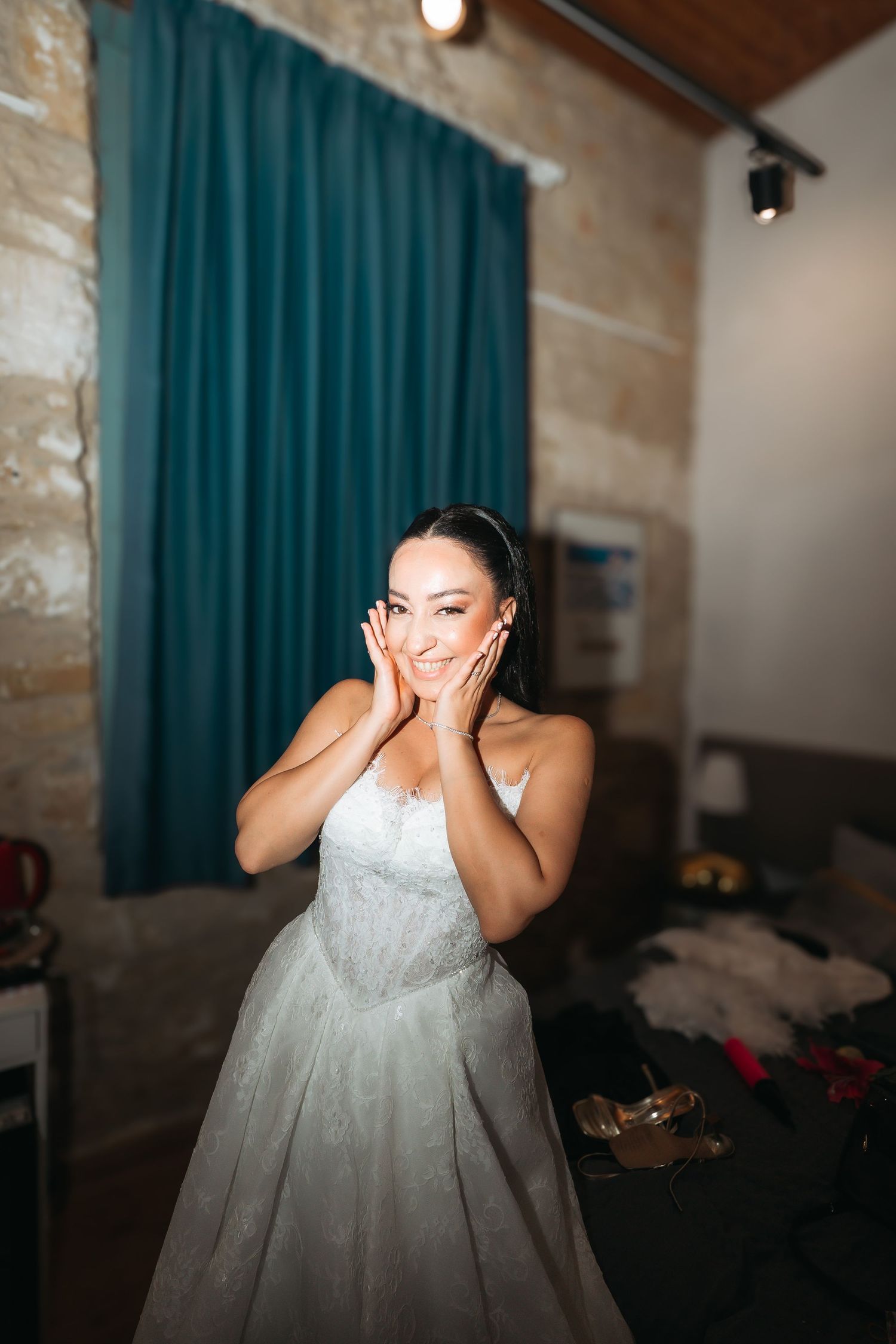 A radiant bride in a white lace gown holds her face with both hands, smiling joyfully in a warmly lit dressing room.