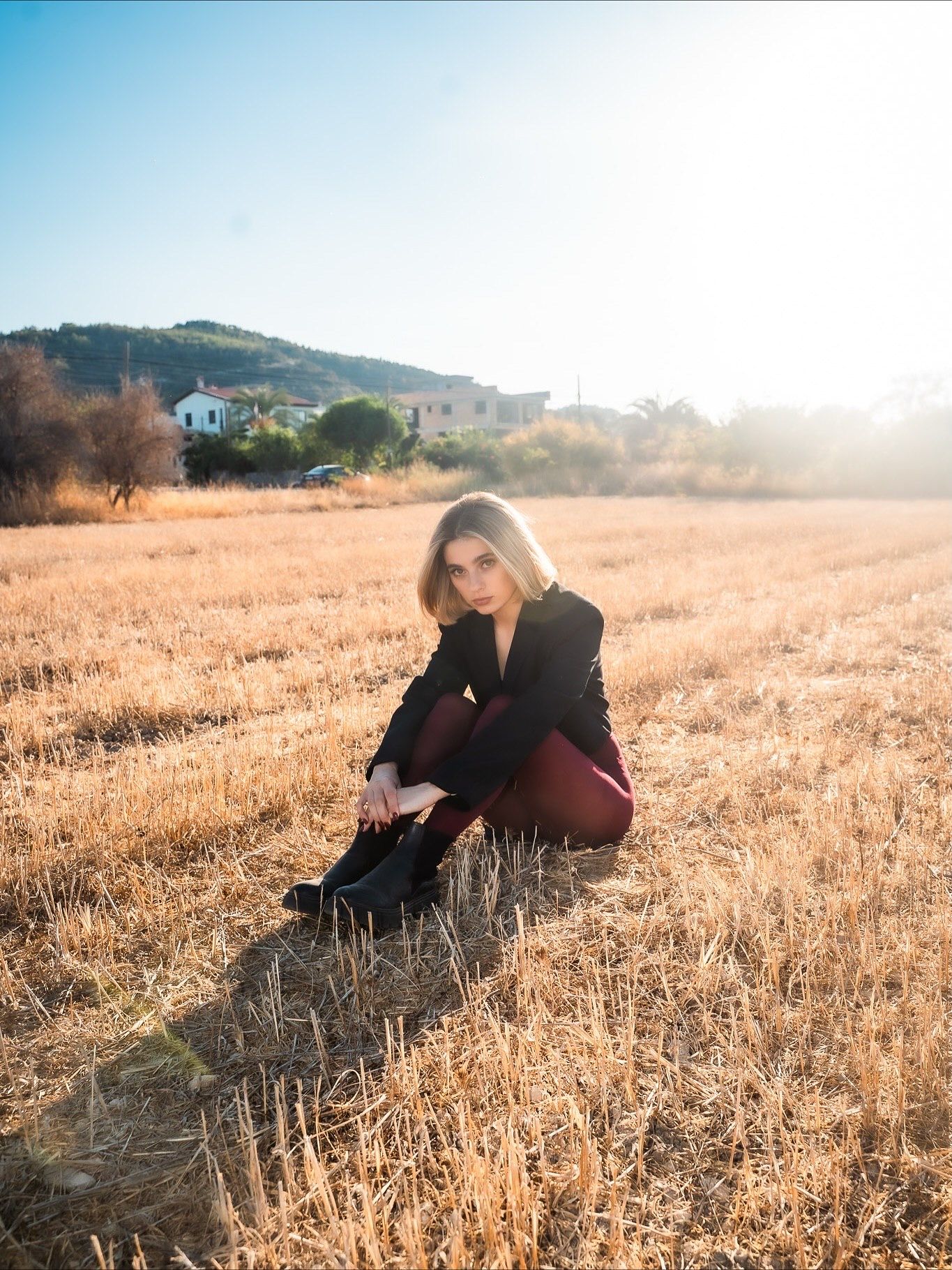 Young woman in black blazer and burgundy pants sitting in a golden field with sunlit hills and buildings behind her.
