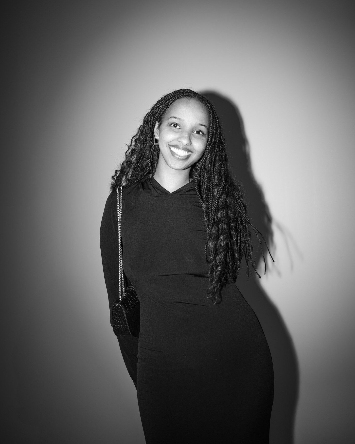 Black and white portrait of a smiling young woman with long braids wearing a black dress, holding a small bag.