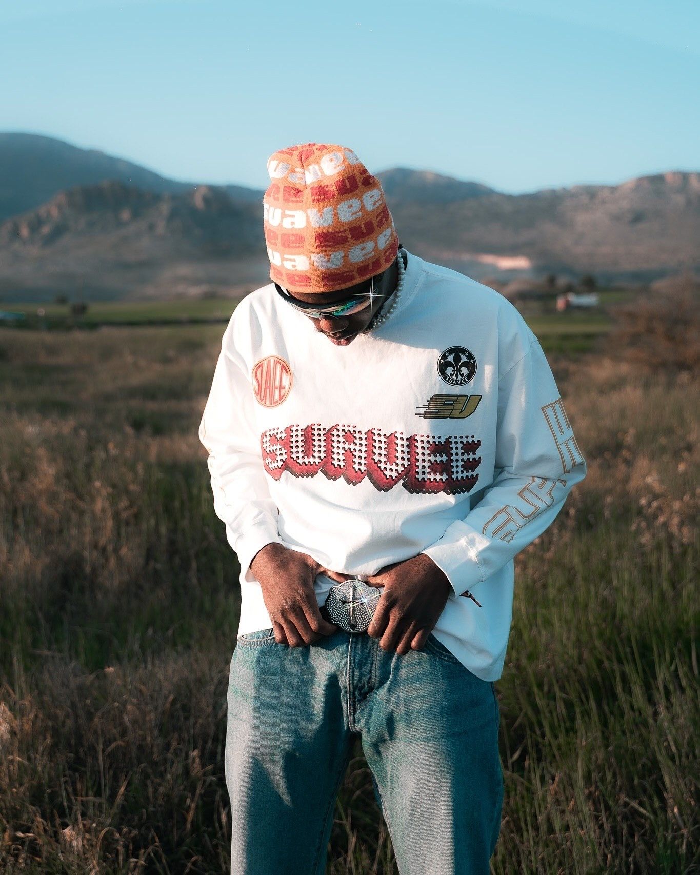 Young man in white graphic sweatshirt, patterned beanie, and jeans standing in a field with mountains behind.