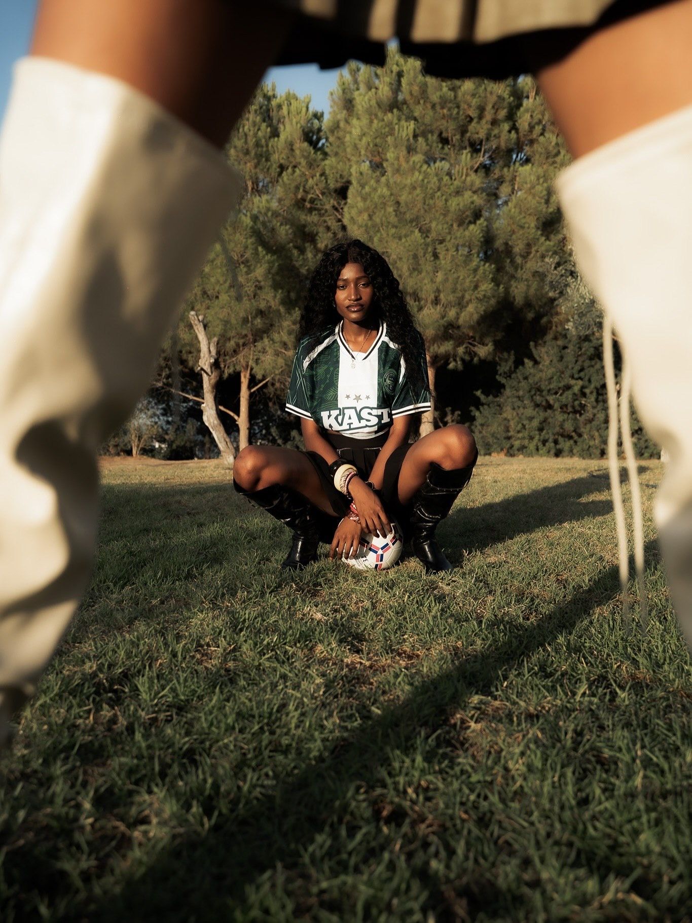 Young woman in green soccer jersey crouching with ball on grass field, framed by white boots in foreground.