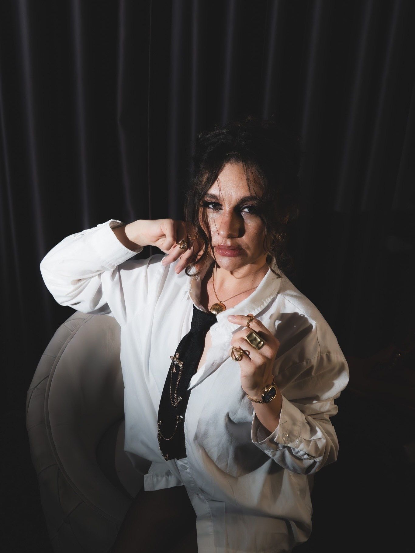 Woman in white shirt with gold rings poses confidently beside white chair in dark studio portrait.