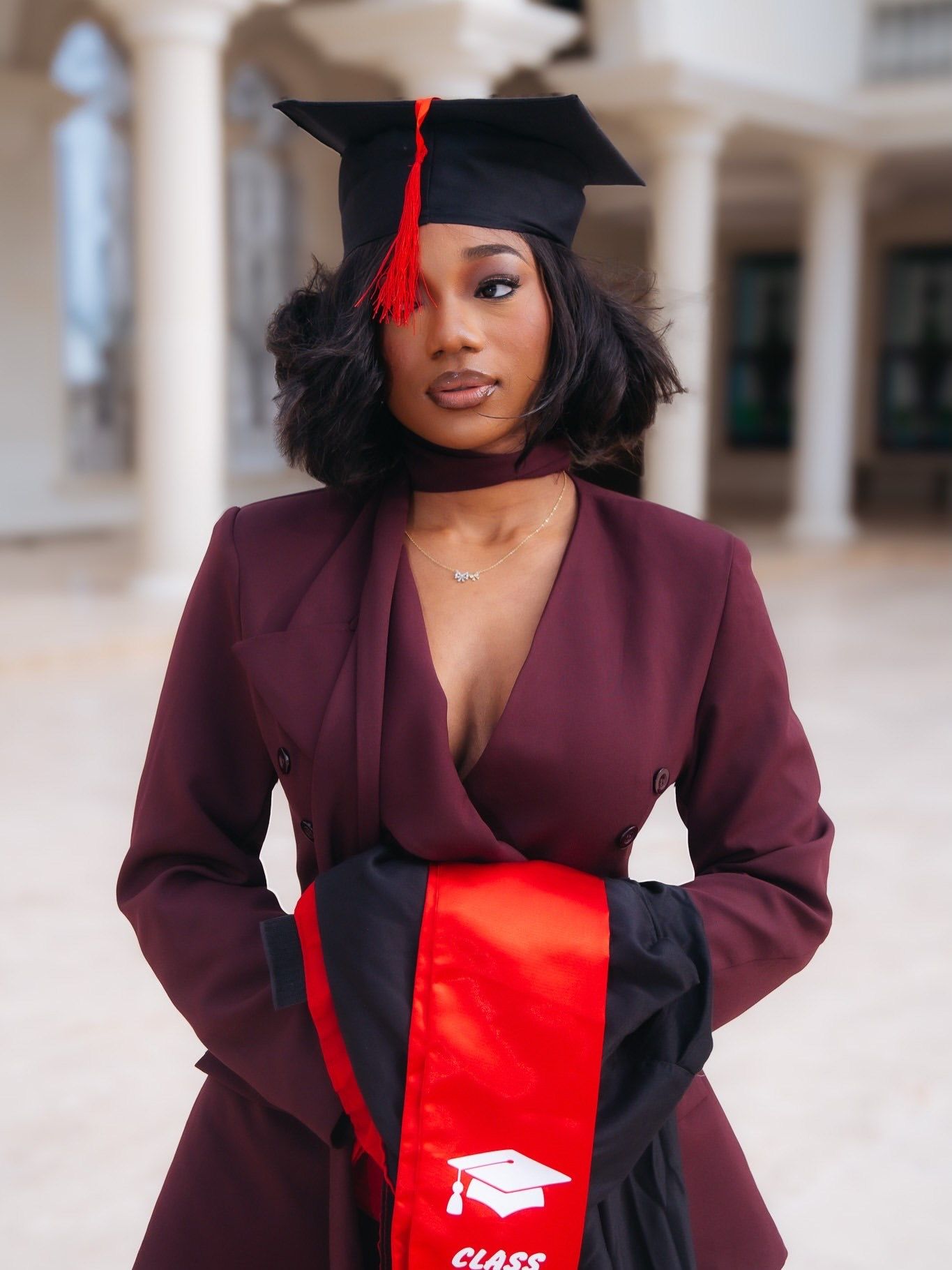 Young woman in maroon blazer suit and graduation cap with red tassel and Class sash poses outdoors.