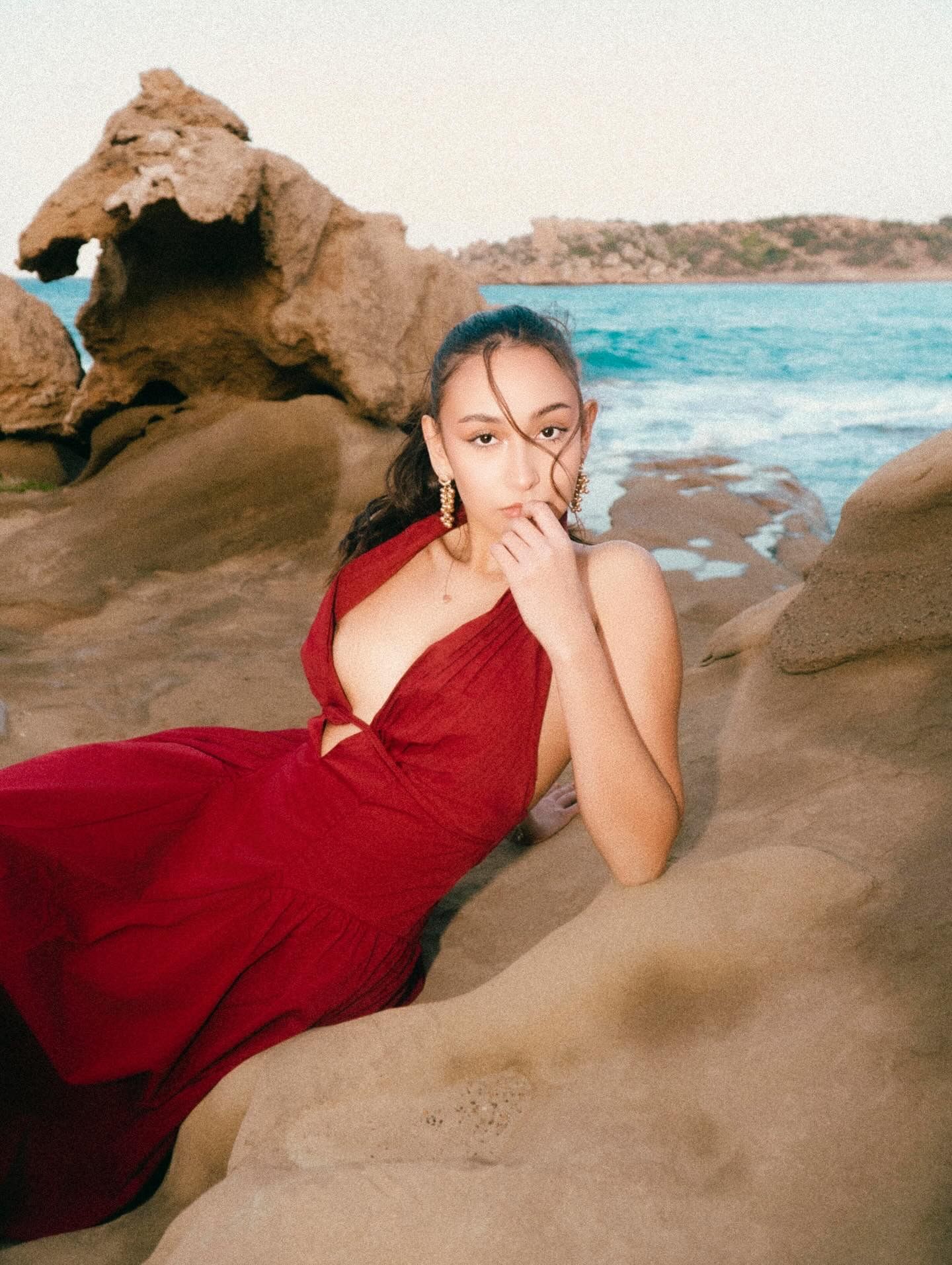 Woman in red dress poses on rocky beach with ocean and hills in background.