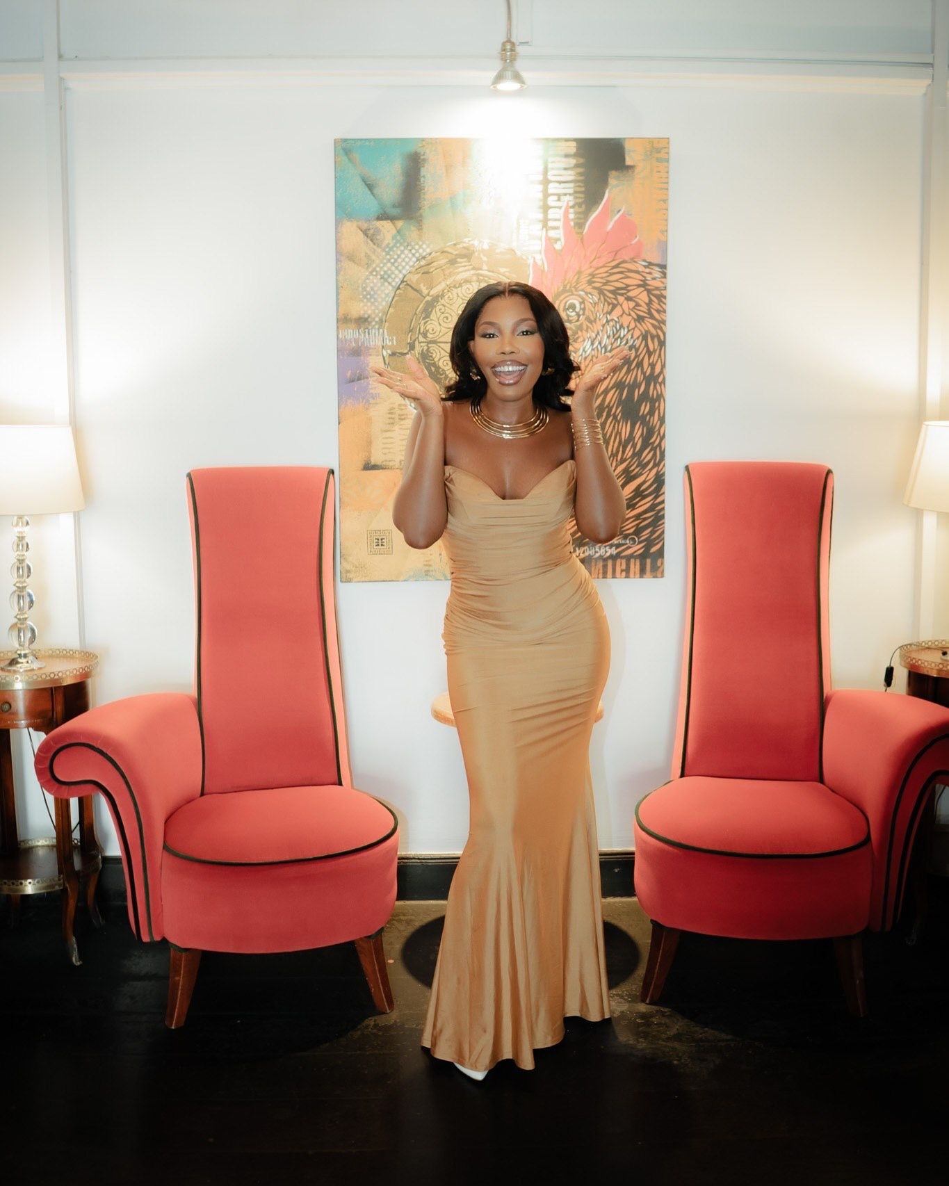 Woman in gold mermaid gown posing between two red high-back chairs with abstract artwork on white wall.