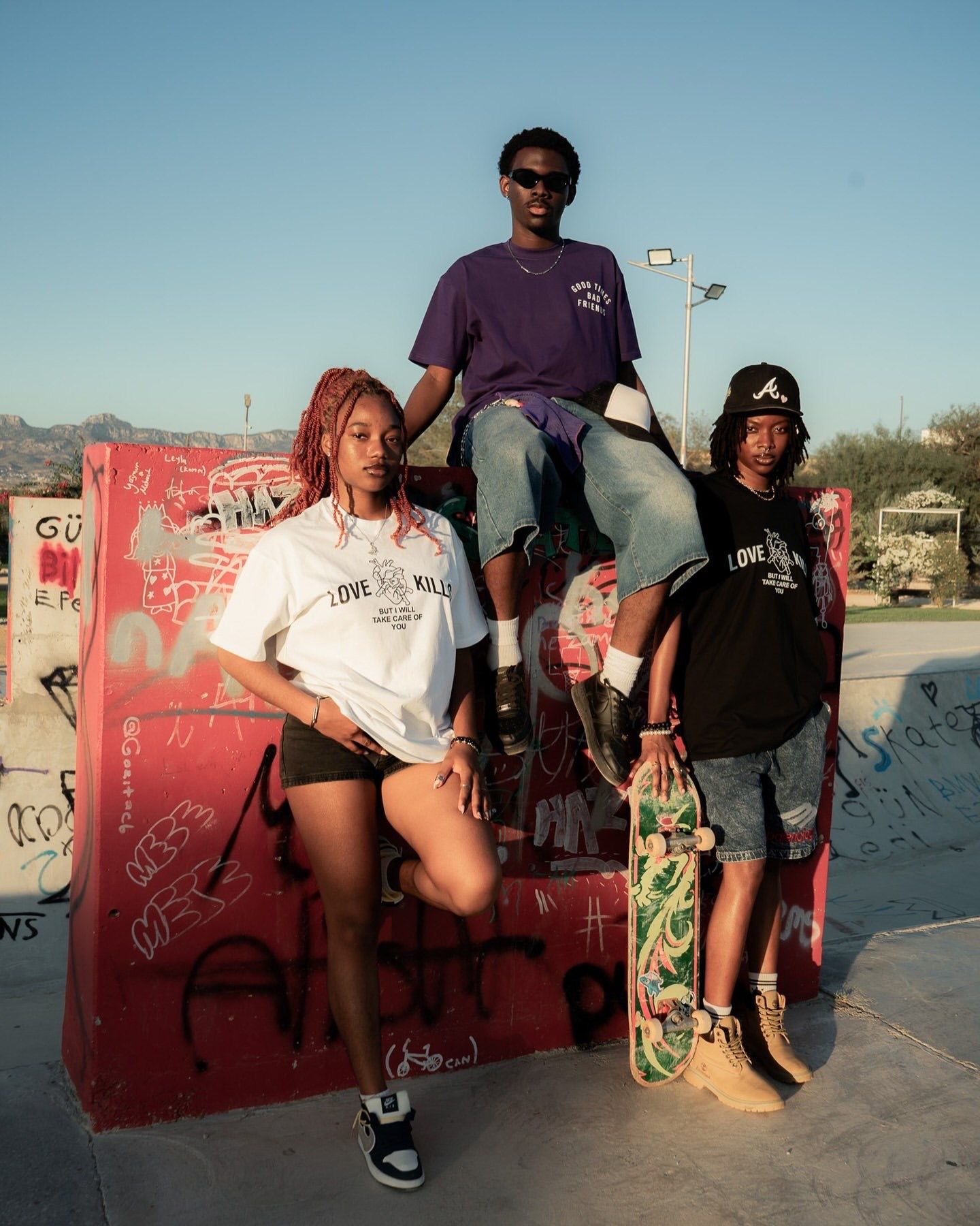 Three stylish young skaters pose at a graffiti-covered skate park, wearing streetwear including purple and white graphic tees.