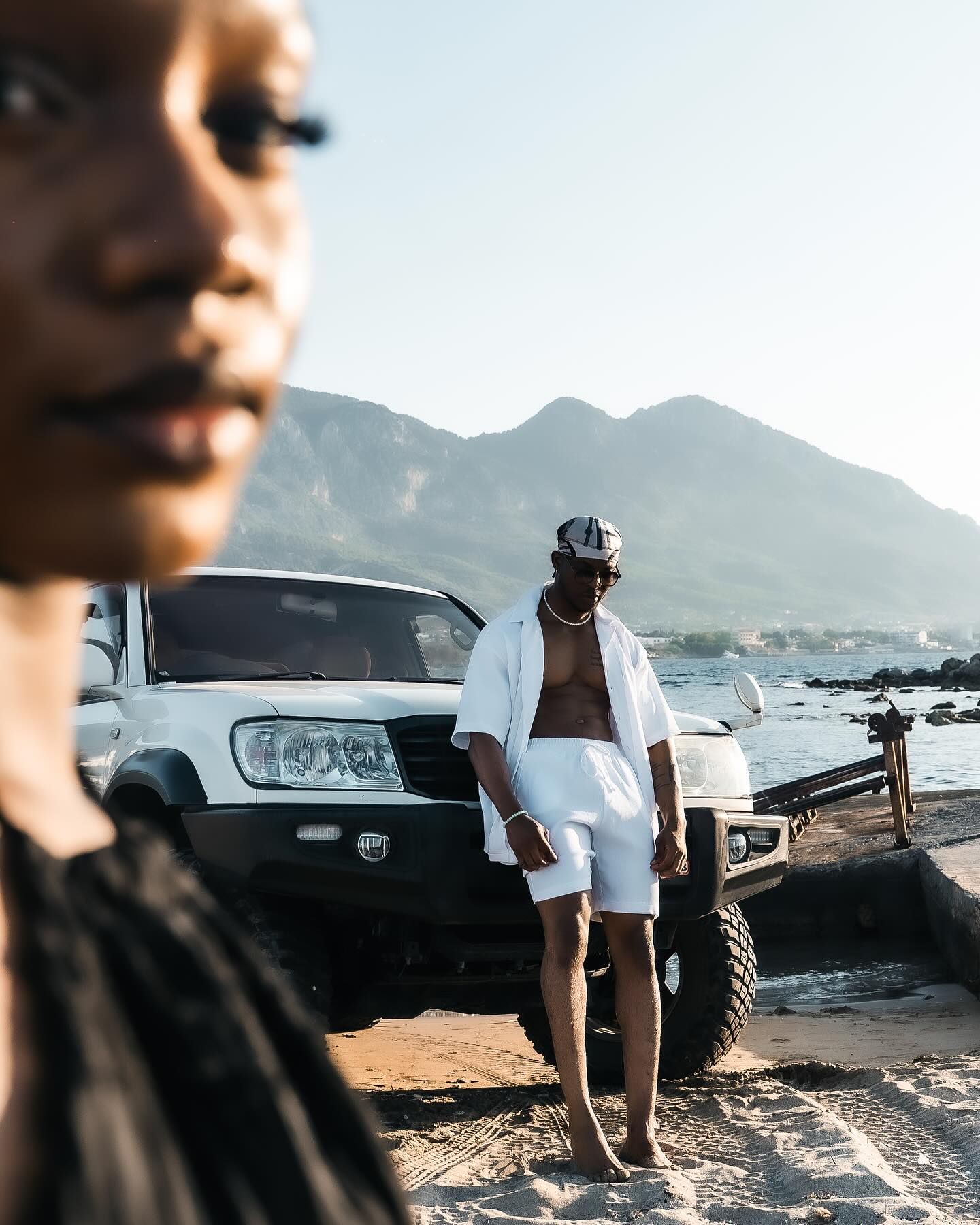 Man in white shorts and open shirt standing by SUV on beach with mountain backdrop.