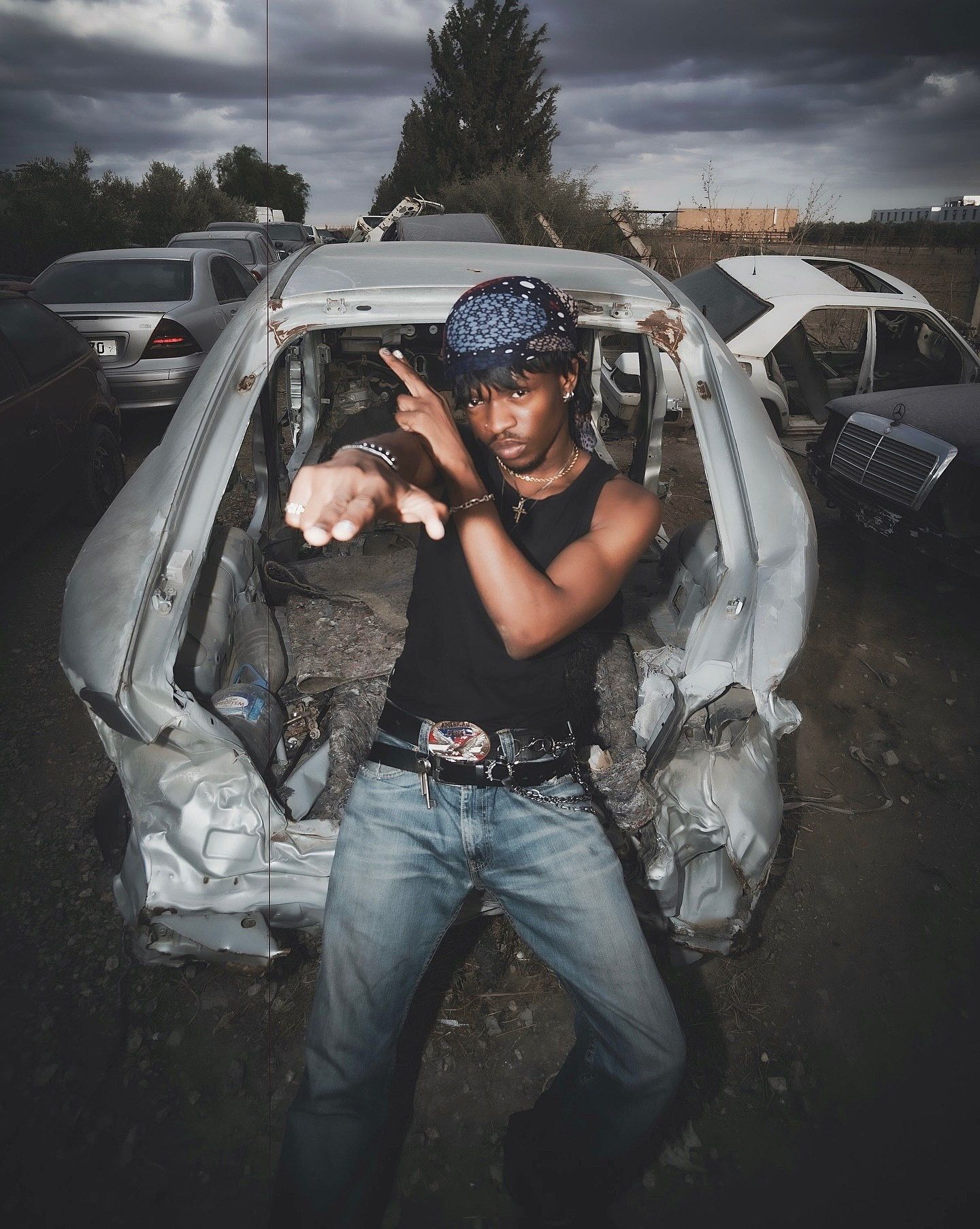 Young man posing confidently in a junkyard surrounded by wrecked cars under a dramatic cloudy sky.