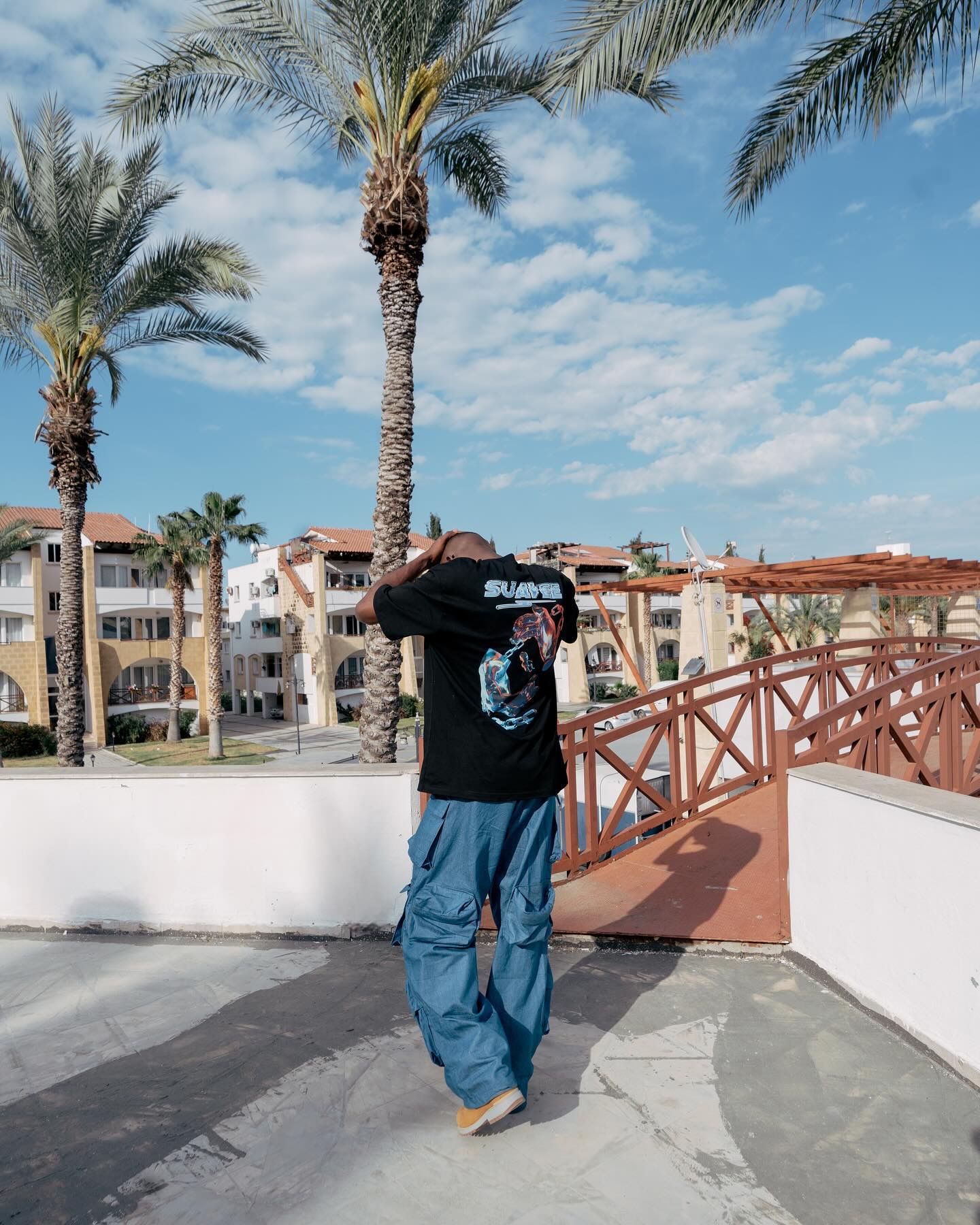 Person in black graphic tee and blue cargo pants standing on rooftop terrace with palm trees and blue sky.