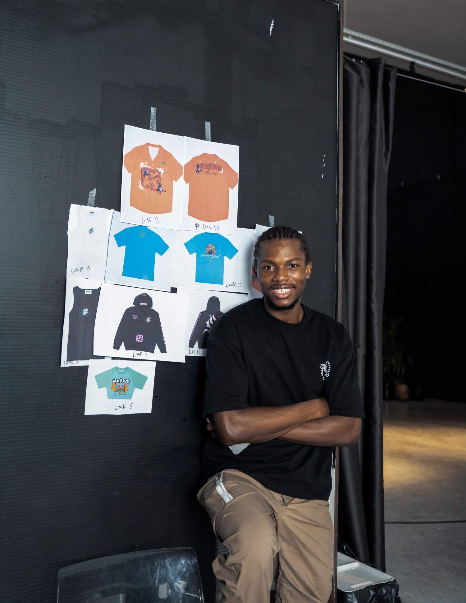 Young man smiling with arms crossed beside pinned clothing design sketches on dark wall.