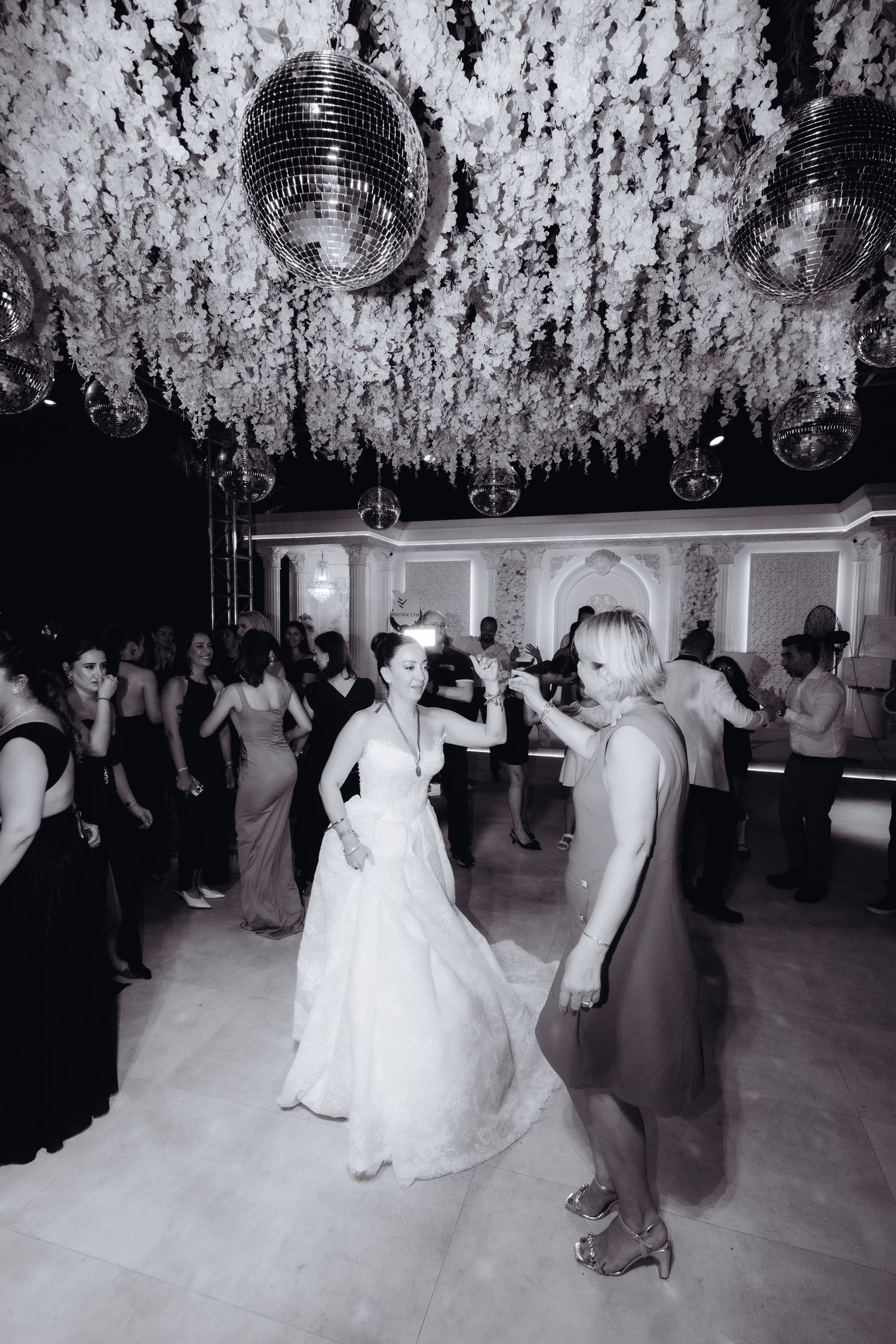 Black and white photo of bride dancing with guest under disco balls and floral ceiling at elegant wedding reception.