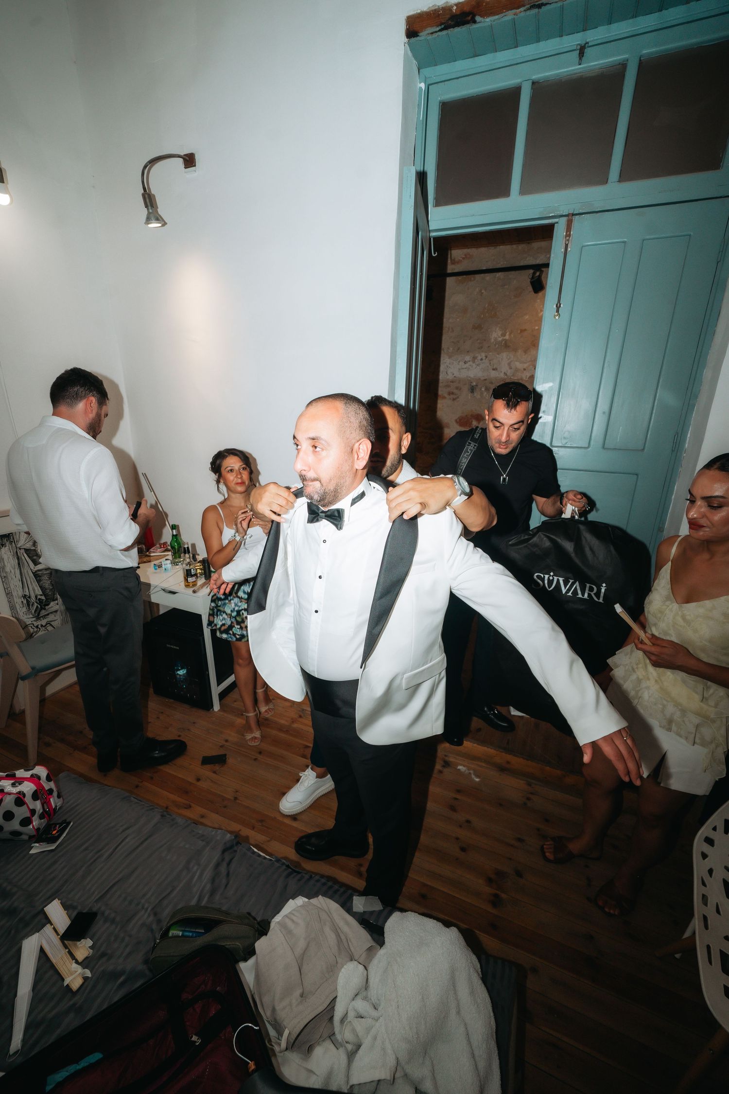 Groom in white tuxedo jacket dancing at wedding reception with guests celebrating in rustic venue.