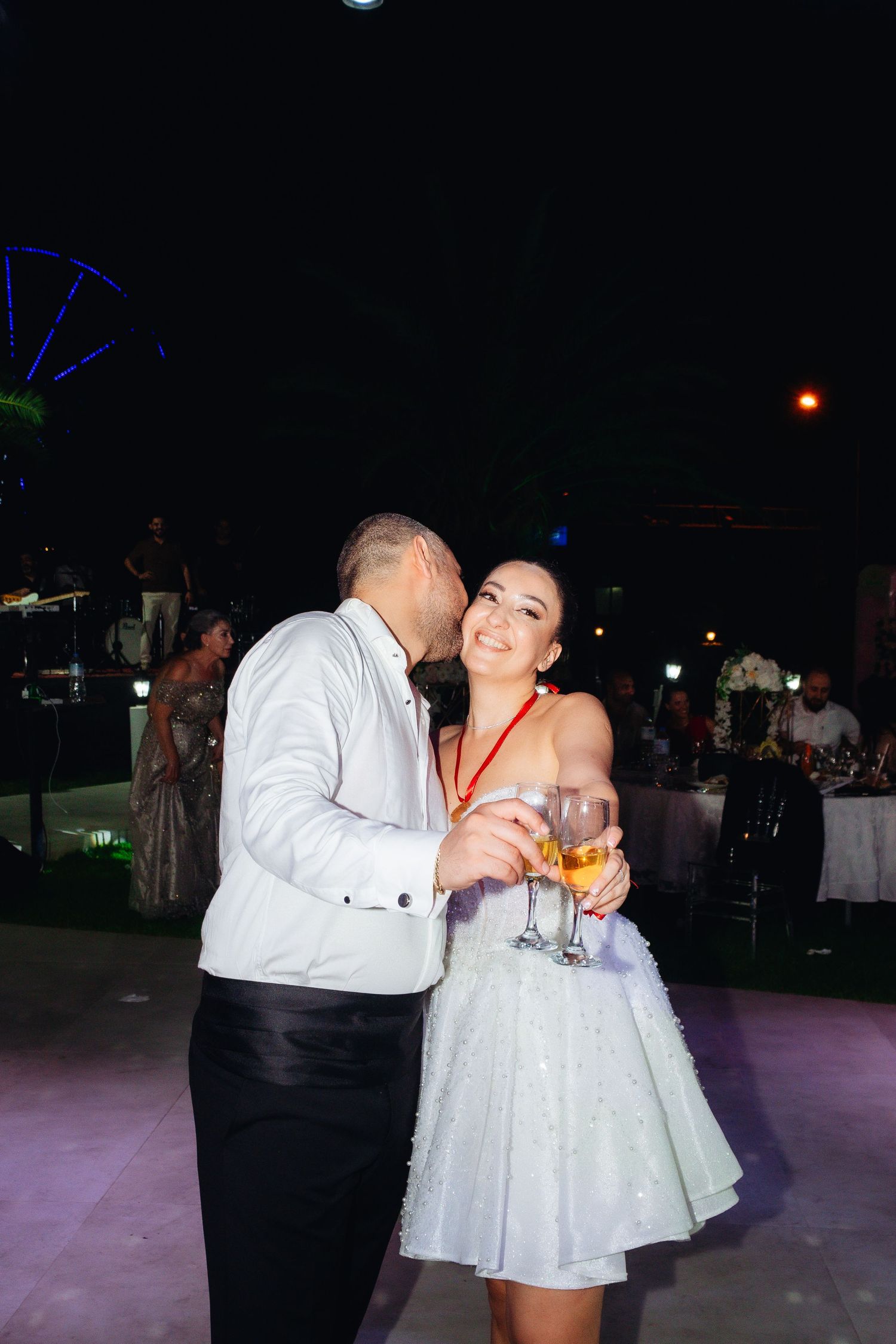 Couple dancing at night event, woman in white dress holding champagne glasses while man whispers in her ear.