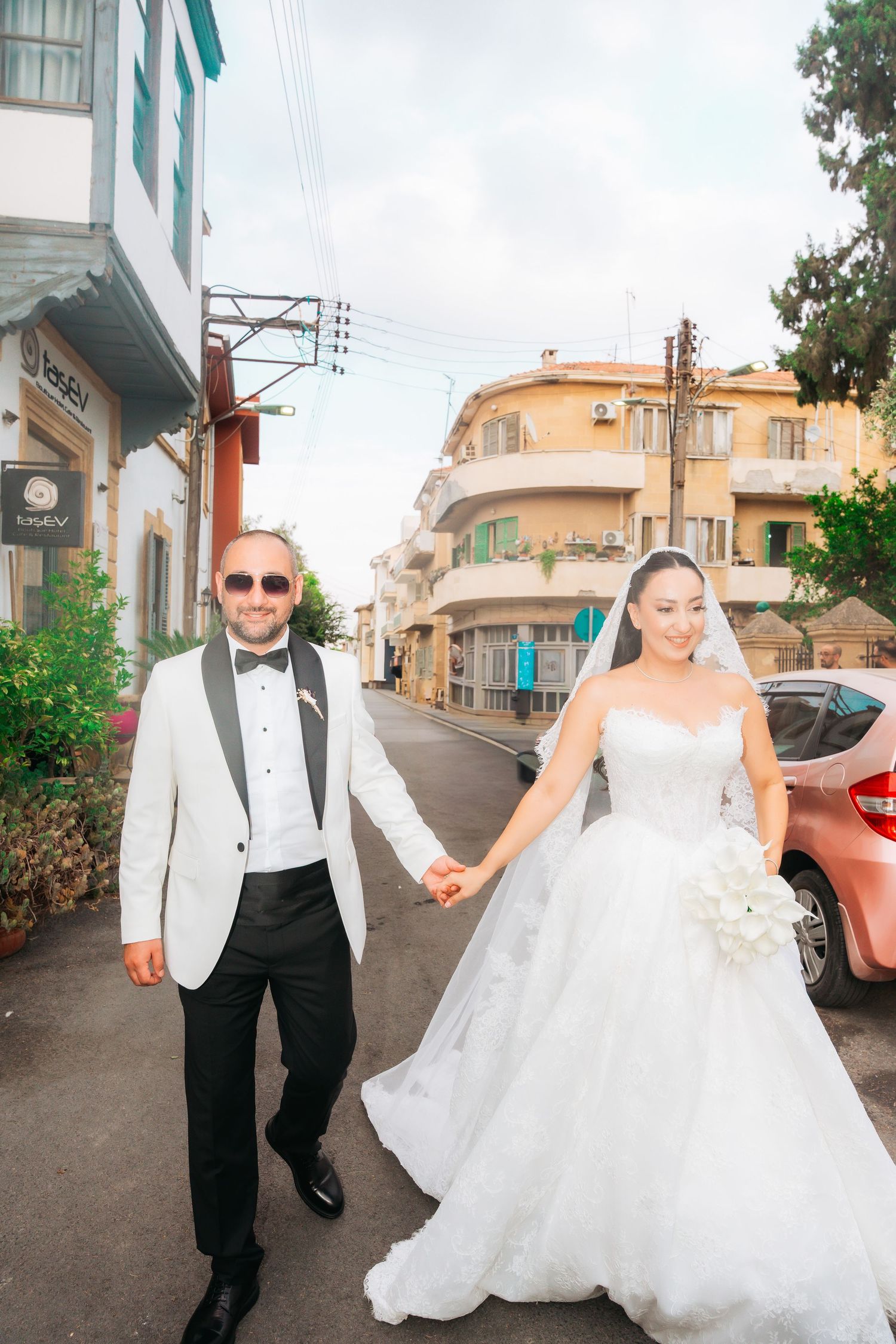 Bride and groom holding hands walking down a sunny street, she in a white gown, he in a white tuxedo jacket.