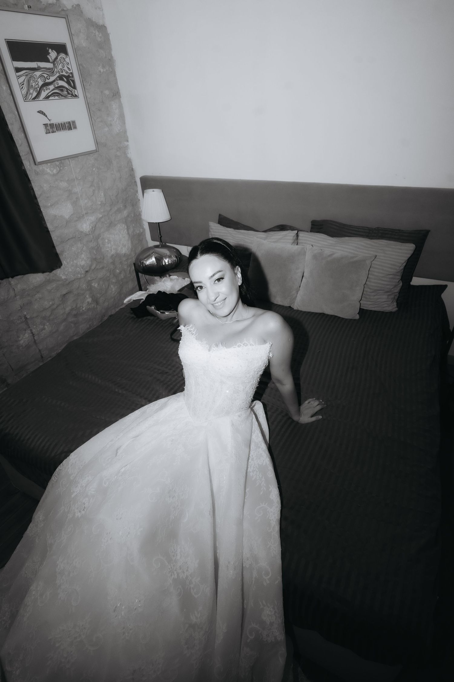 Black and white photo of a smiling bride in a white ball gown sitting on a bed in an elegant hotel room.