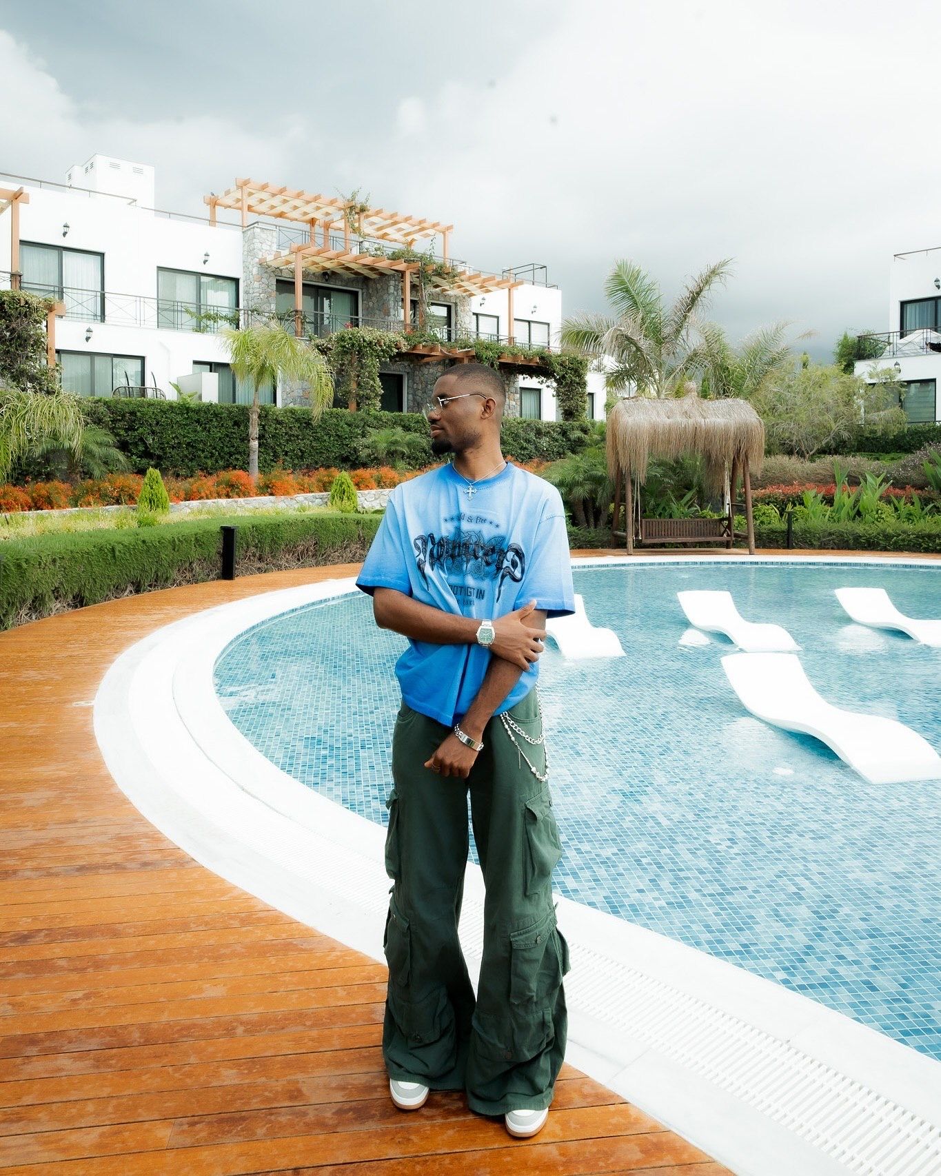Young man in blue graphic tee and cargo pants standing by a resort pool with white loungers and lush greenery.