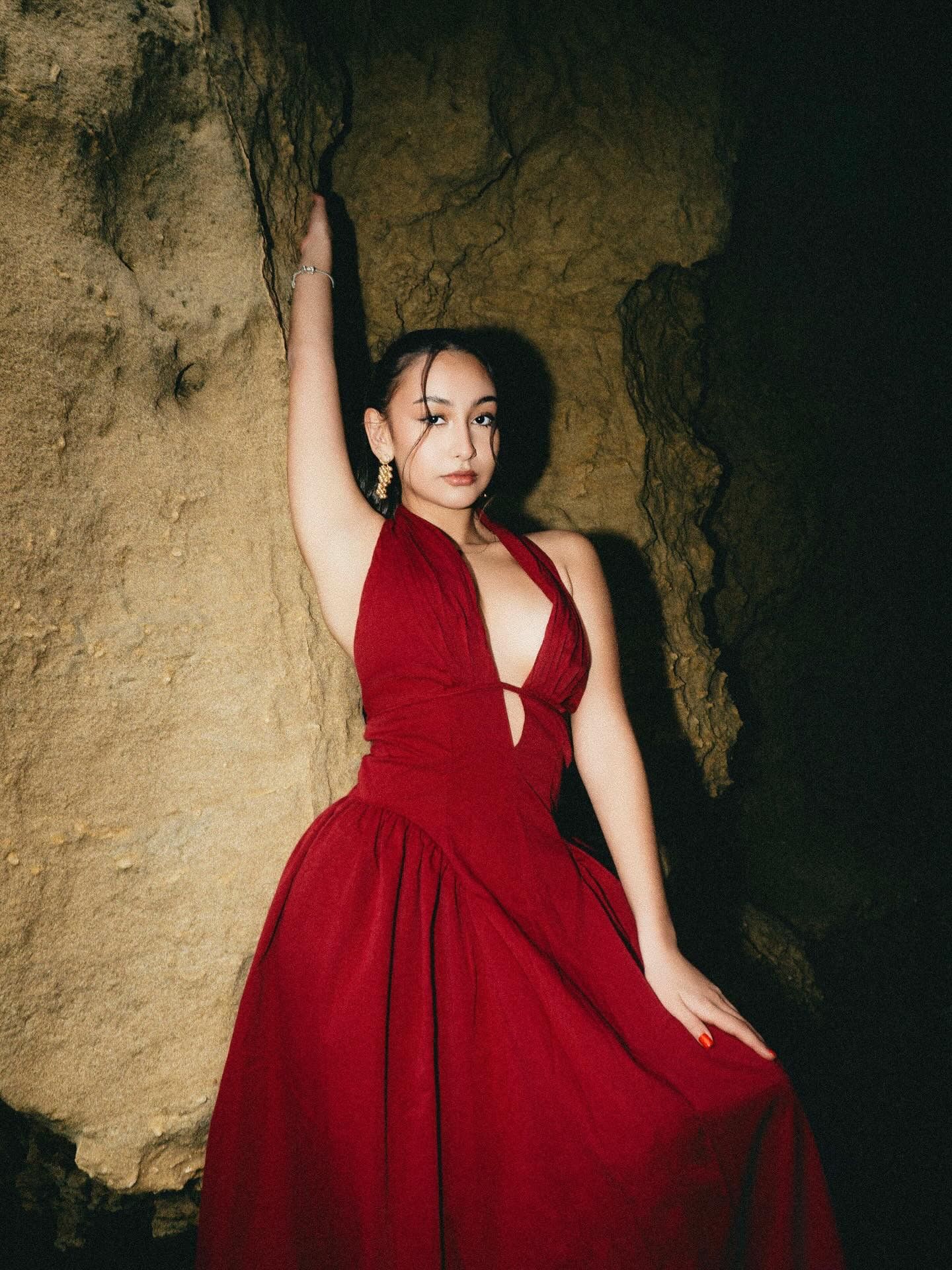 Woman in red halter dress poses dramatically against rocky cave wall in moody editorial photo.