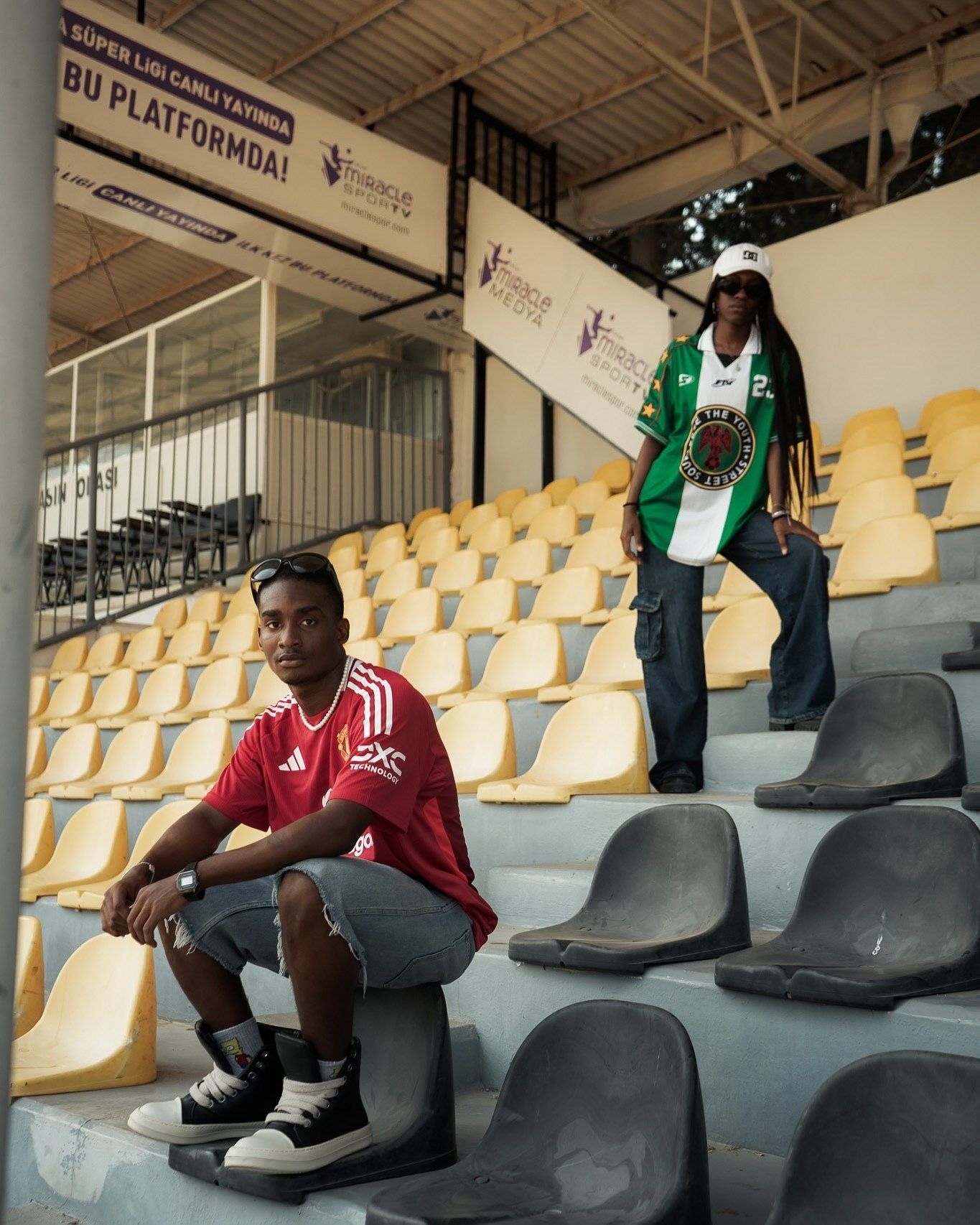 Two young men in sports jerseys pose in yellow and gray stadium seats at a soccer arena.