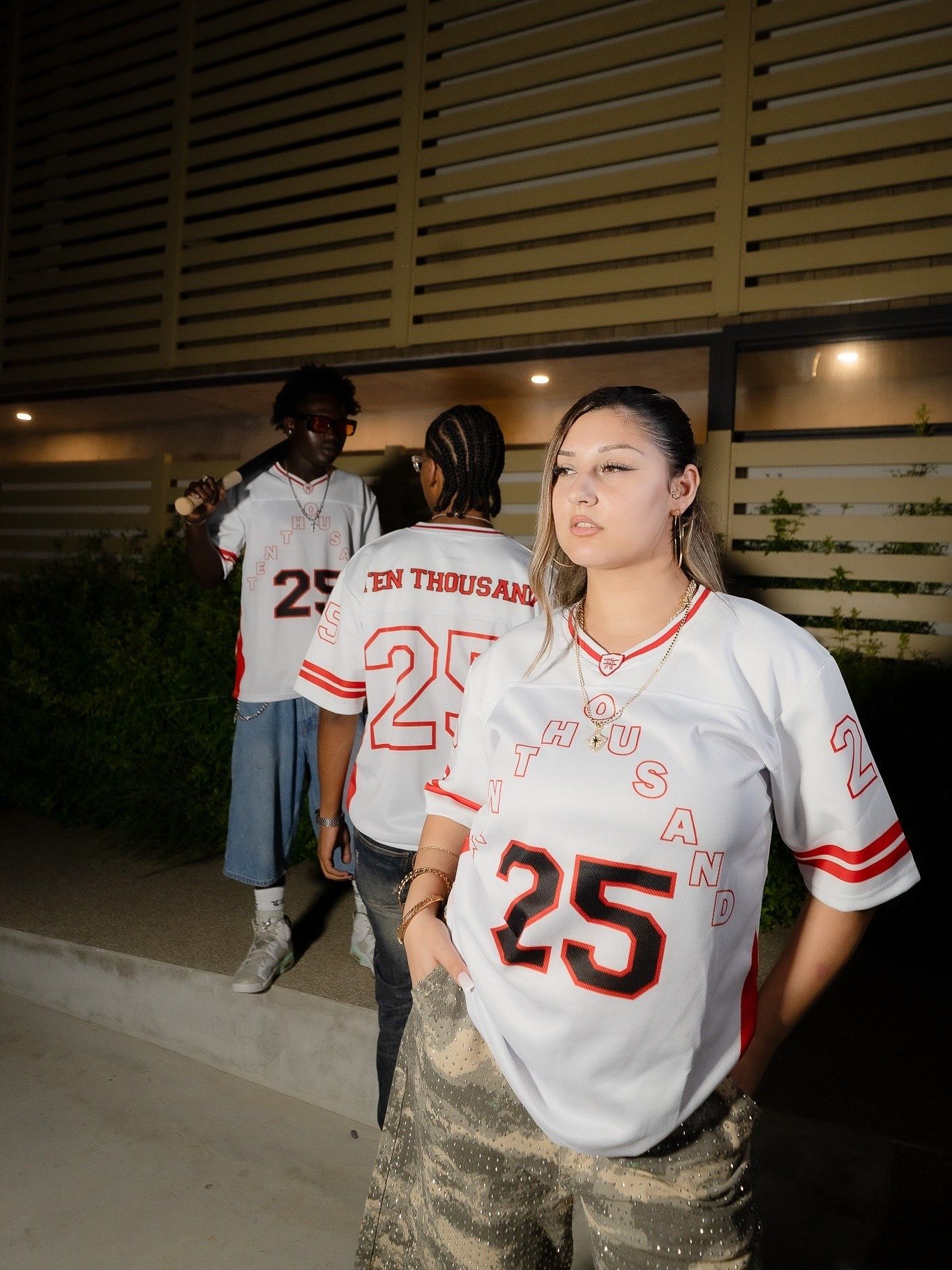 Three people wearing matching white number 25 Ten Thousand jerseys stand outside a building at night.