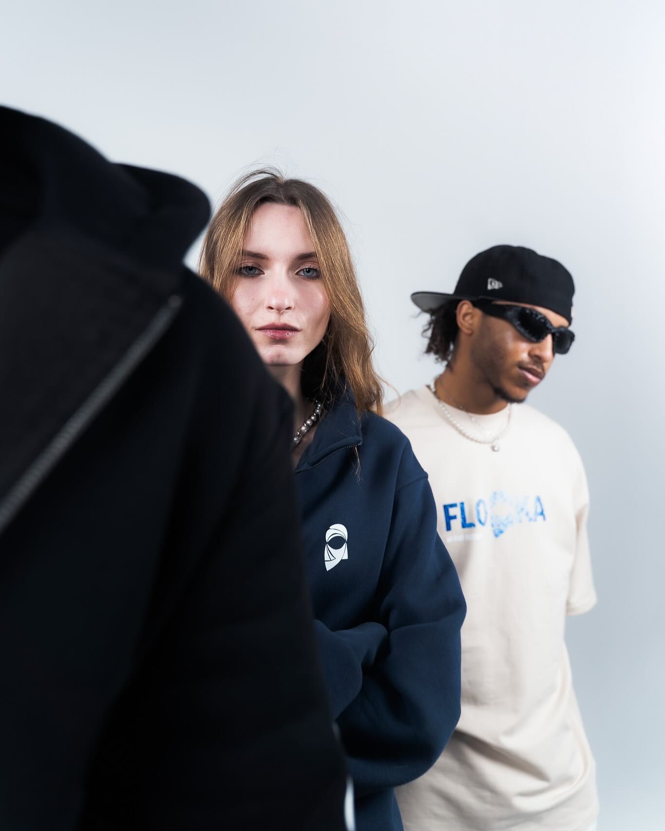 Two young people in streetwear, woman in navy hoodie and man in cream Florida tee and black cap, posing against grey background.