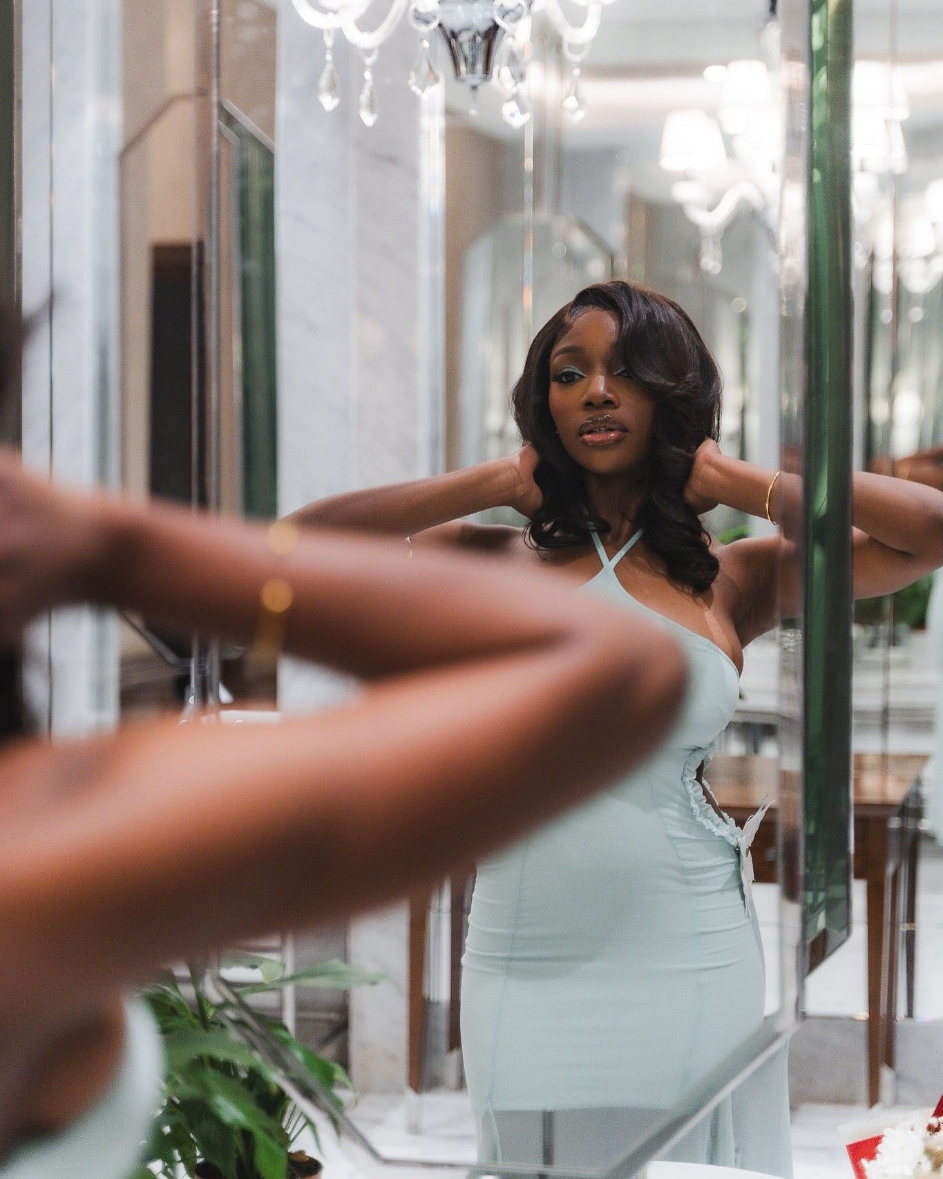 Woman in mint halter dress poses in elegant mirrored bathroom with chandelier overhead.