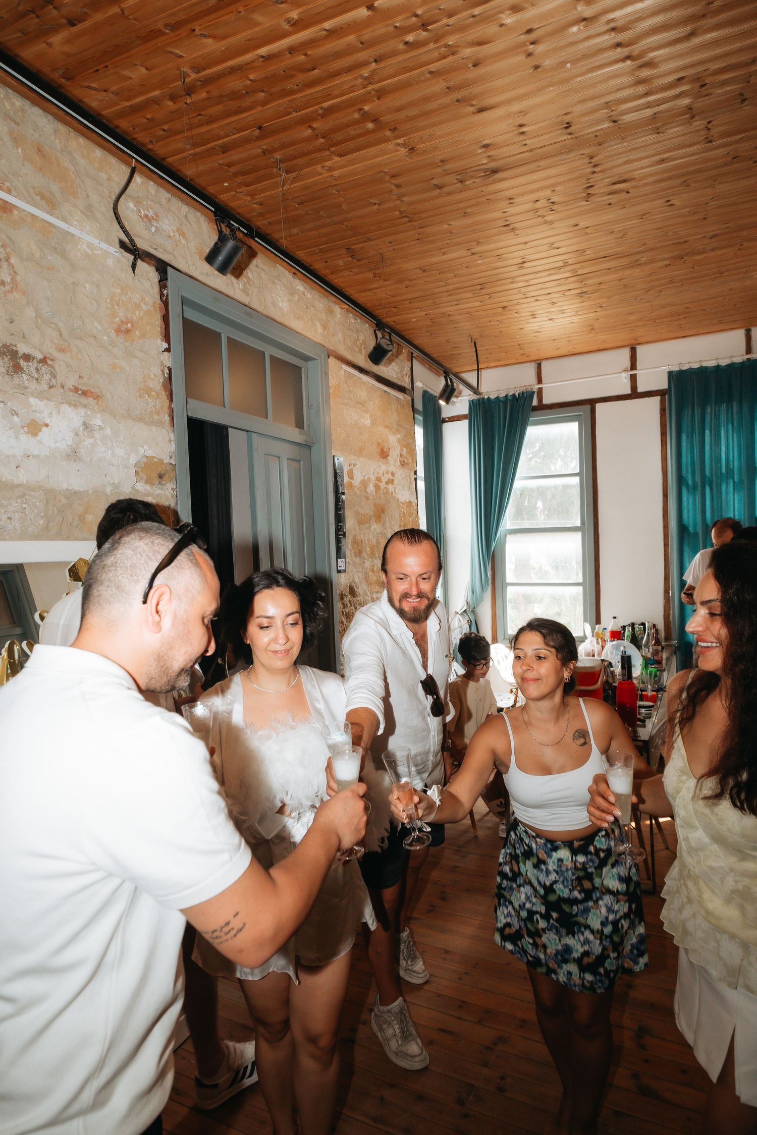 Group of friends in white outfits celebrating indoors, toasting with drinks in a rustic venue with wooden ceiling.