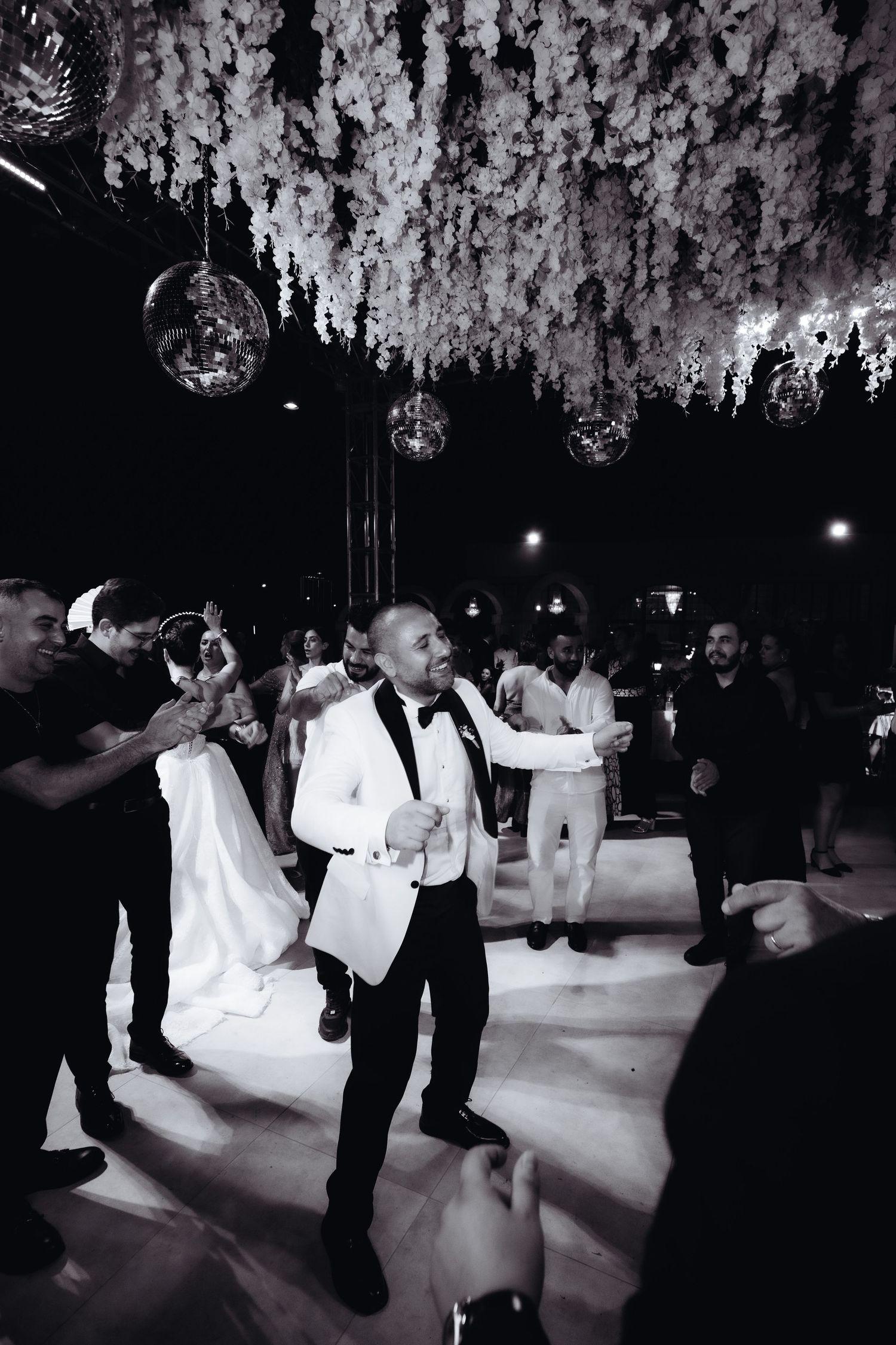 Black and white wedding reception photo with groom in white tuxedo dancing under floral ceiling decorations.