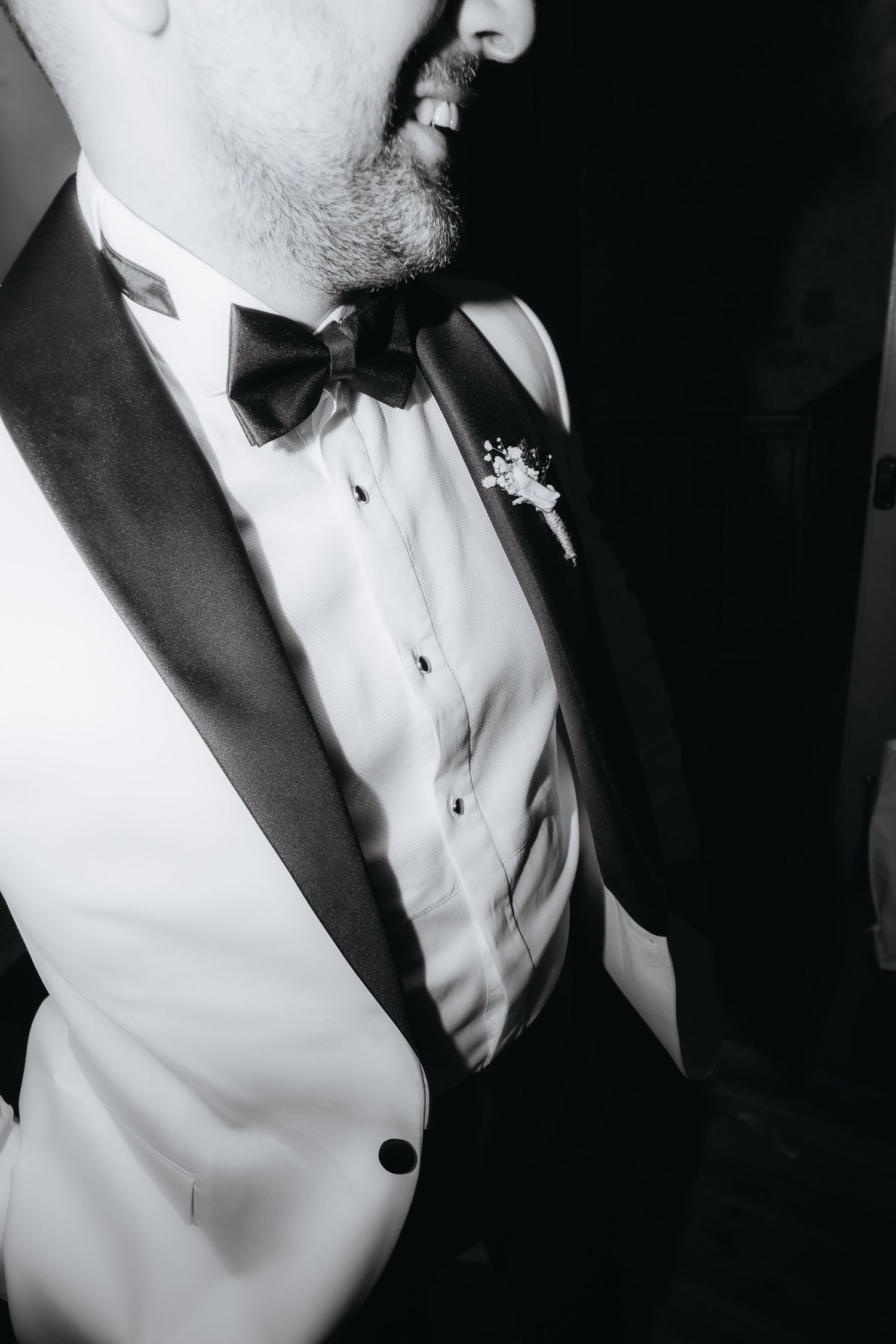 Black and white photo of a groom in a tuxedo with bow tie and boutonniere, smiling elegantly.