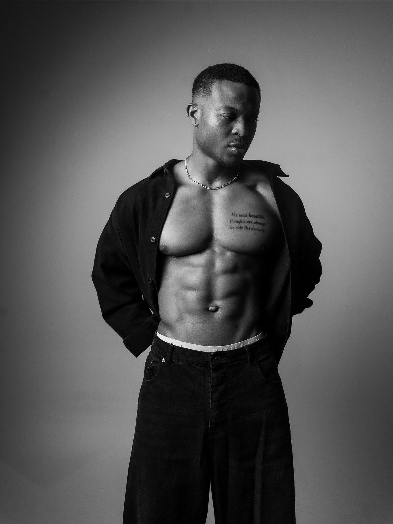 Black and white photo of a muscular man with open shirt showing defined abs and a chest tattoo, posing confidently.