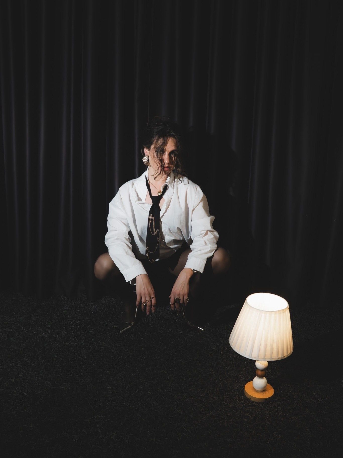 Person in white shirt crouches on dark floor beside small glowing lamp in dramatic low-light photo.