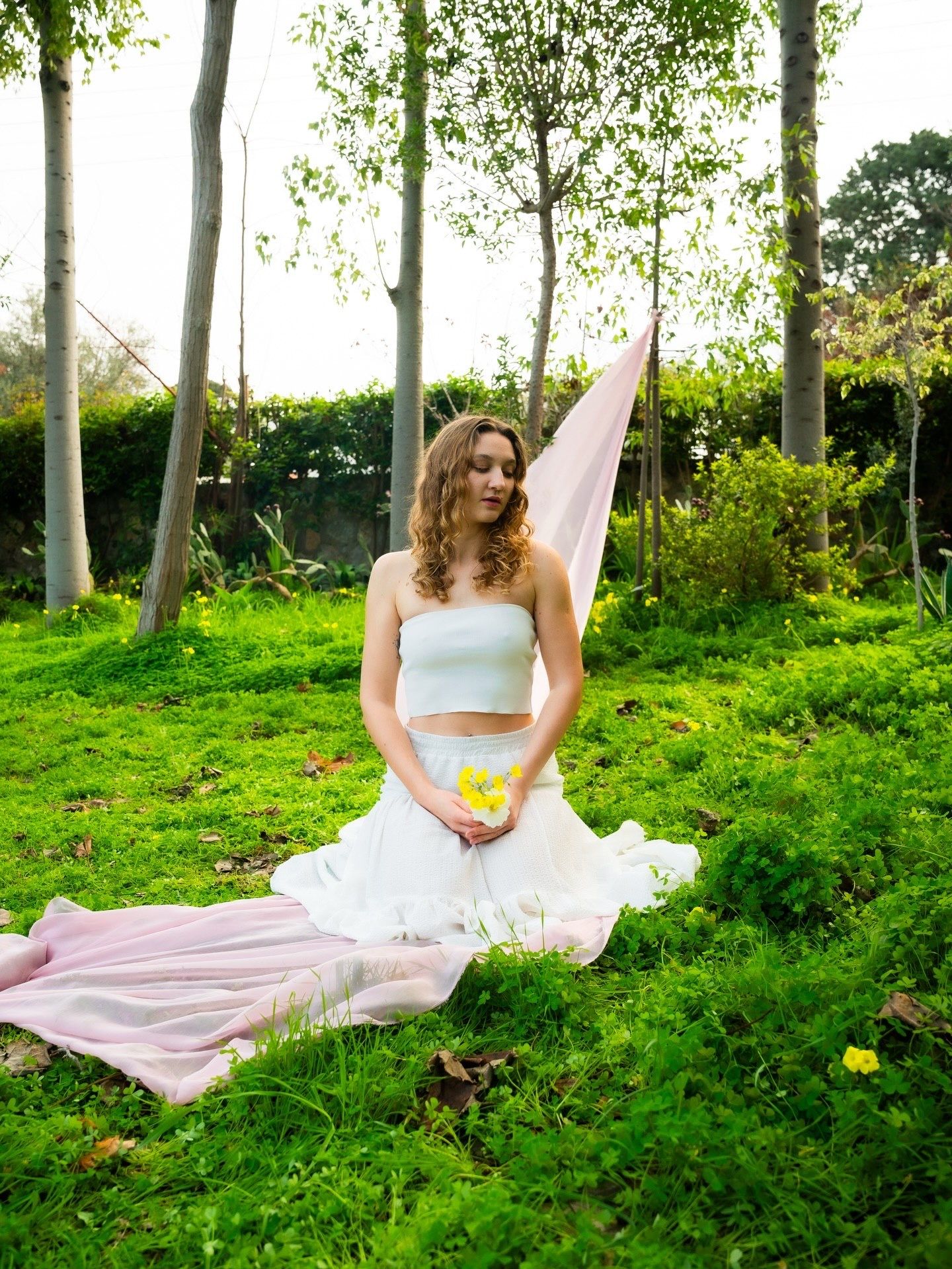 Young woman in white skirt and crop top sits on lush green grass holding yellow flowers with pink fabric behind her.