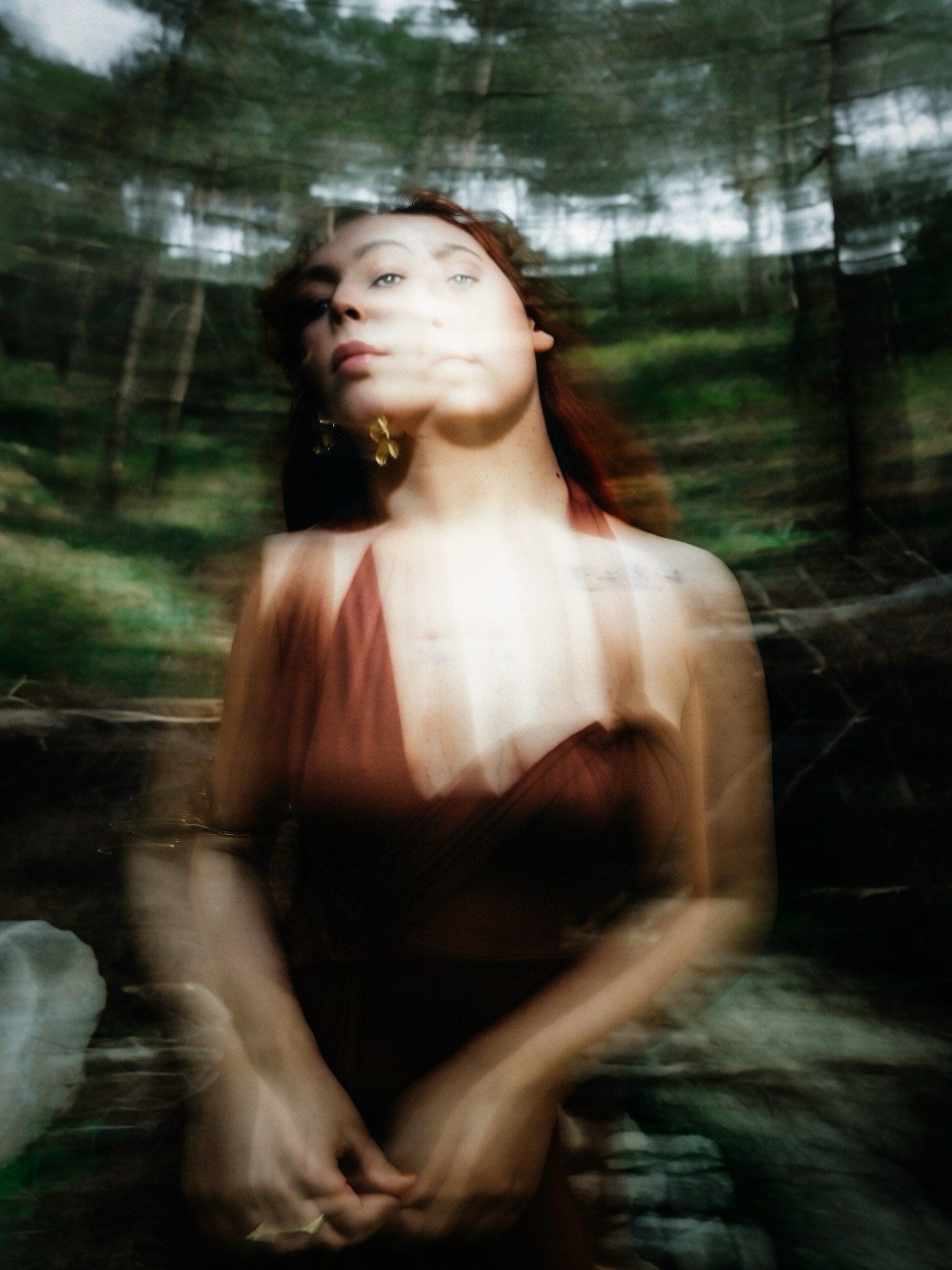 Artistic double-exposure portrait of a woman in red dress surrounded by blurred forest greenery.