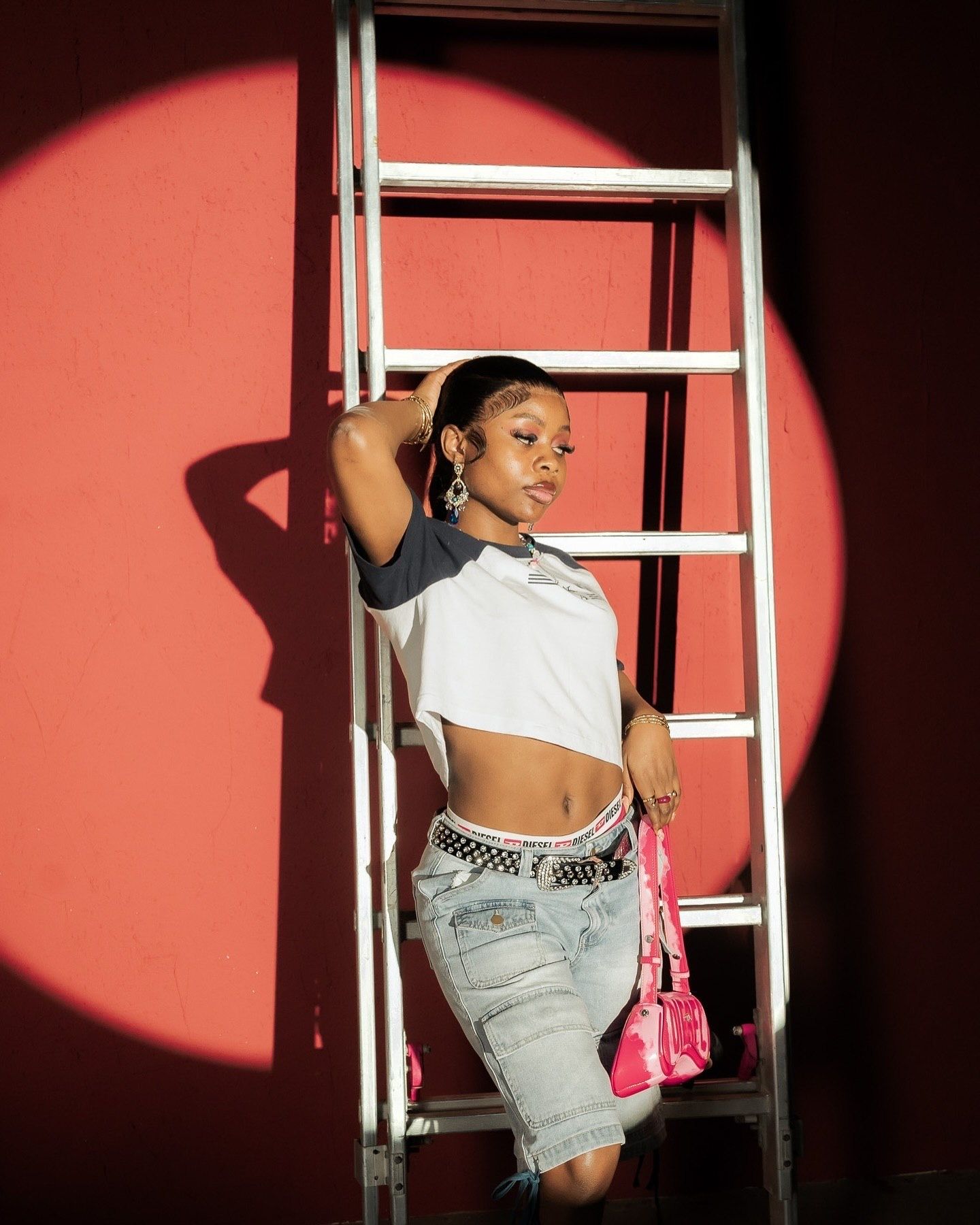 Young woman in white crop top and denim shorts poses against red wall with white ladder in dramatic spotlight.