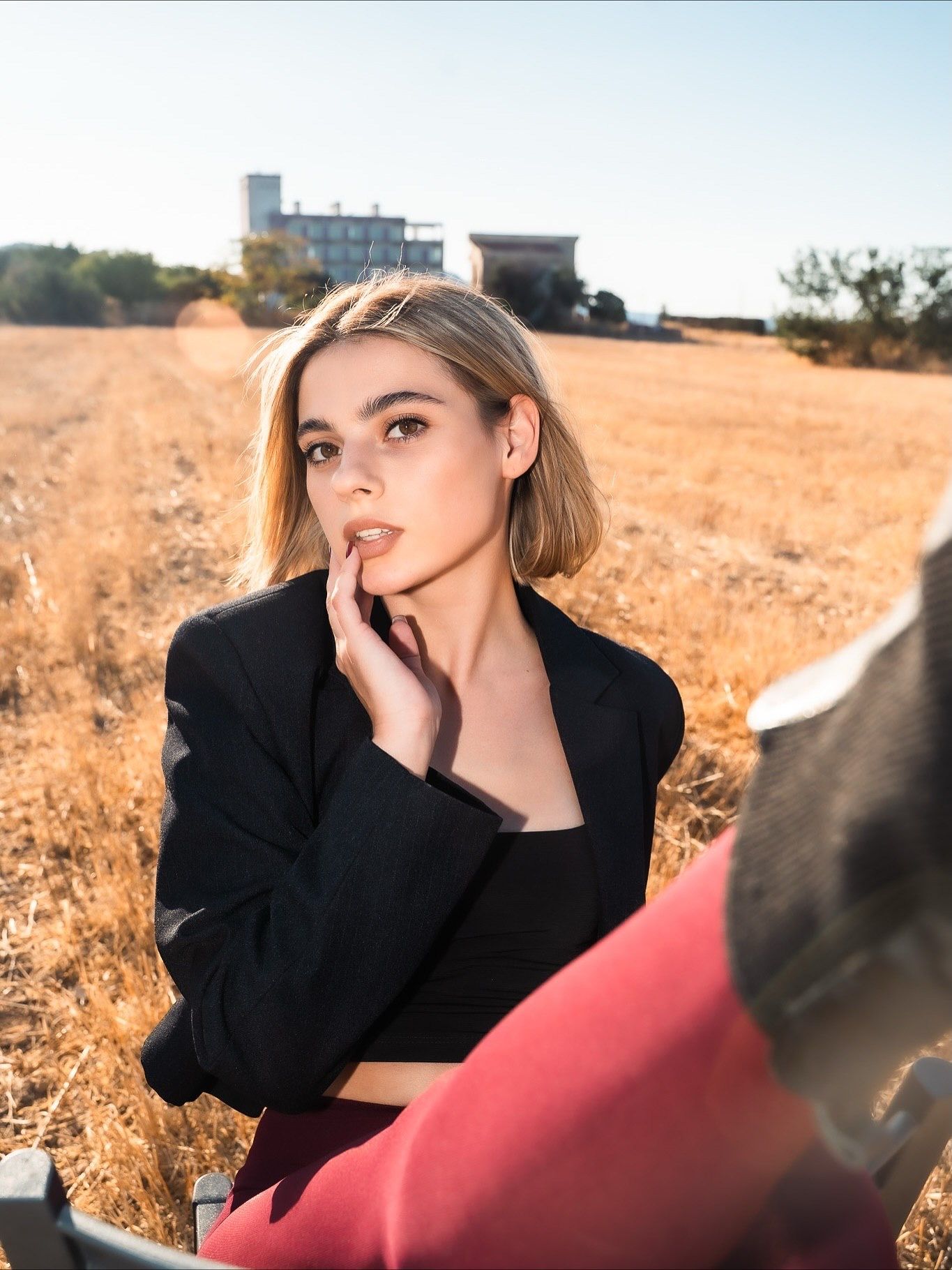 Young blonde woman in black blazer and red pants poses thoughtfully in golden field with building in background.