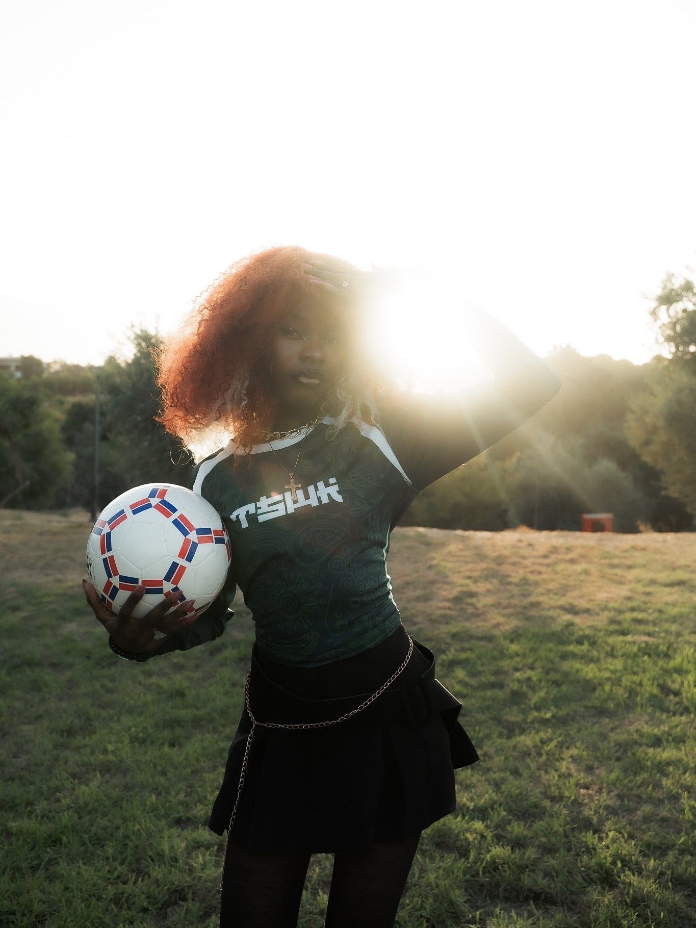 Woman with curly red hair holding a soccer ball outdoors at golden hour in a dark sports jersey.