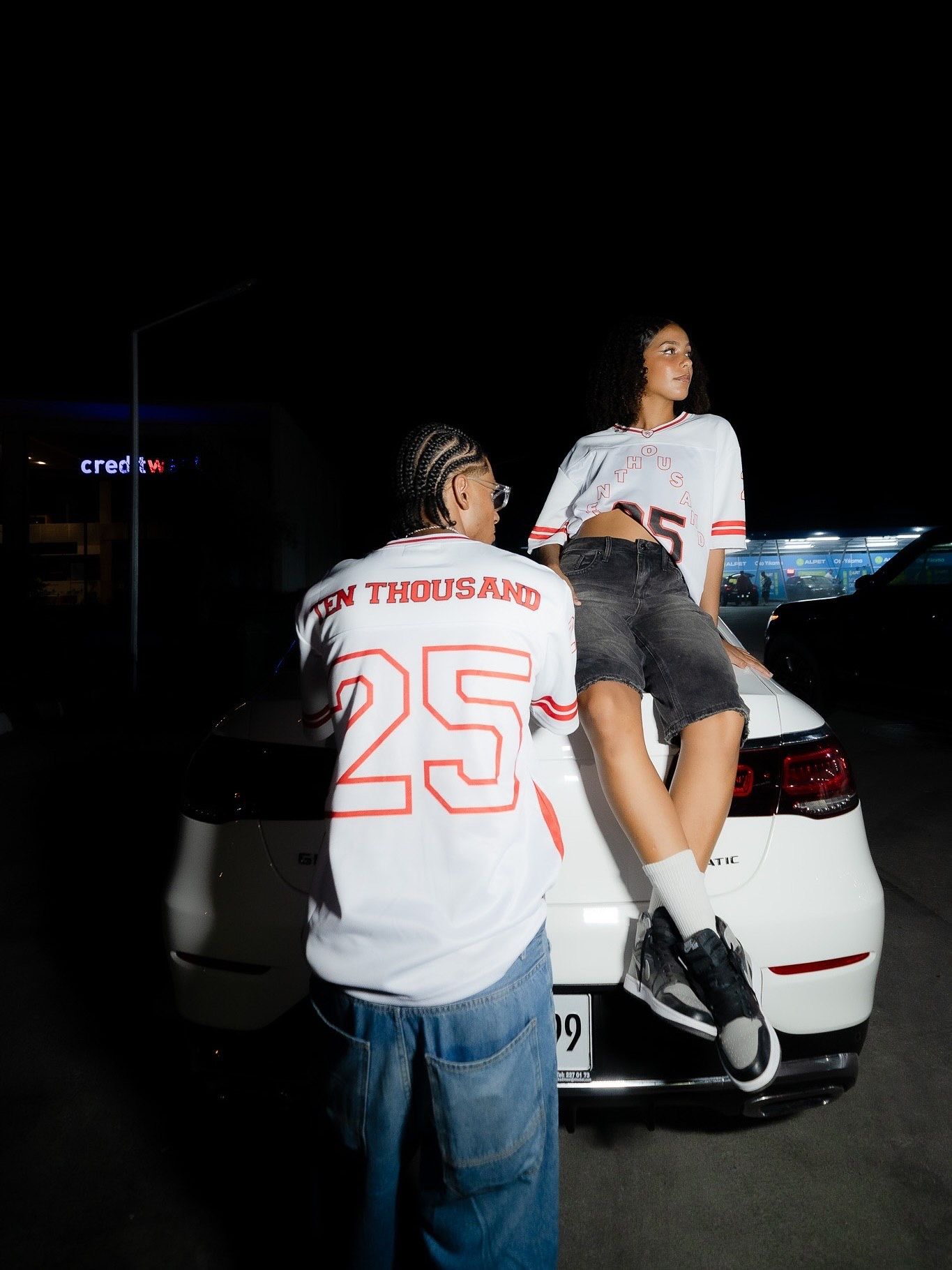 Two people wearing matching white number 25 jerseys pose on a white car at night.