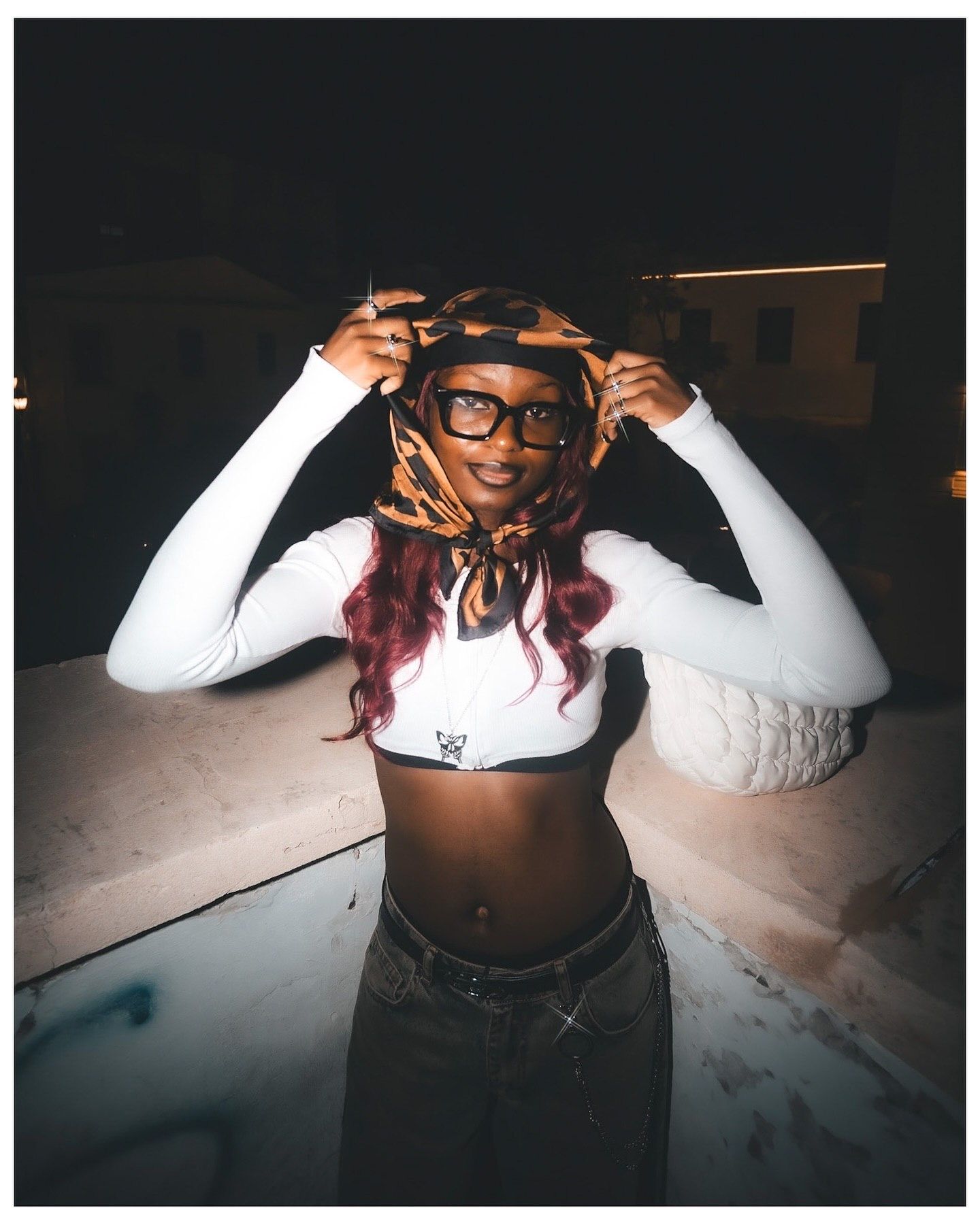 Young woman in white crop top and glasses poses outdoors at night with colorful headscarf and red hair.