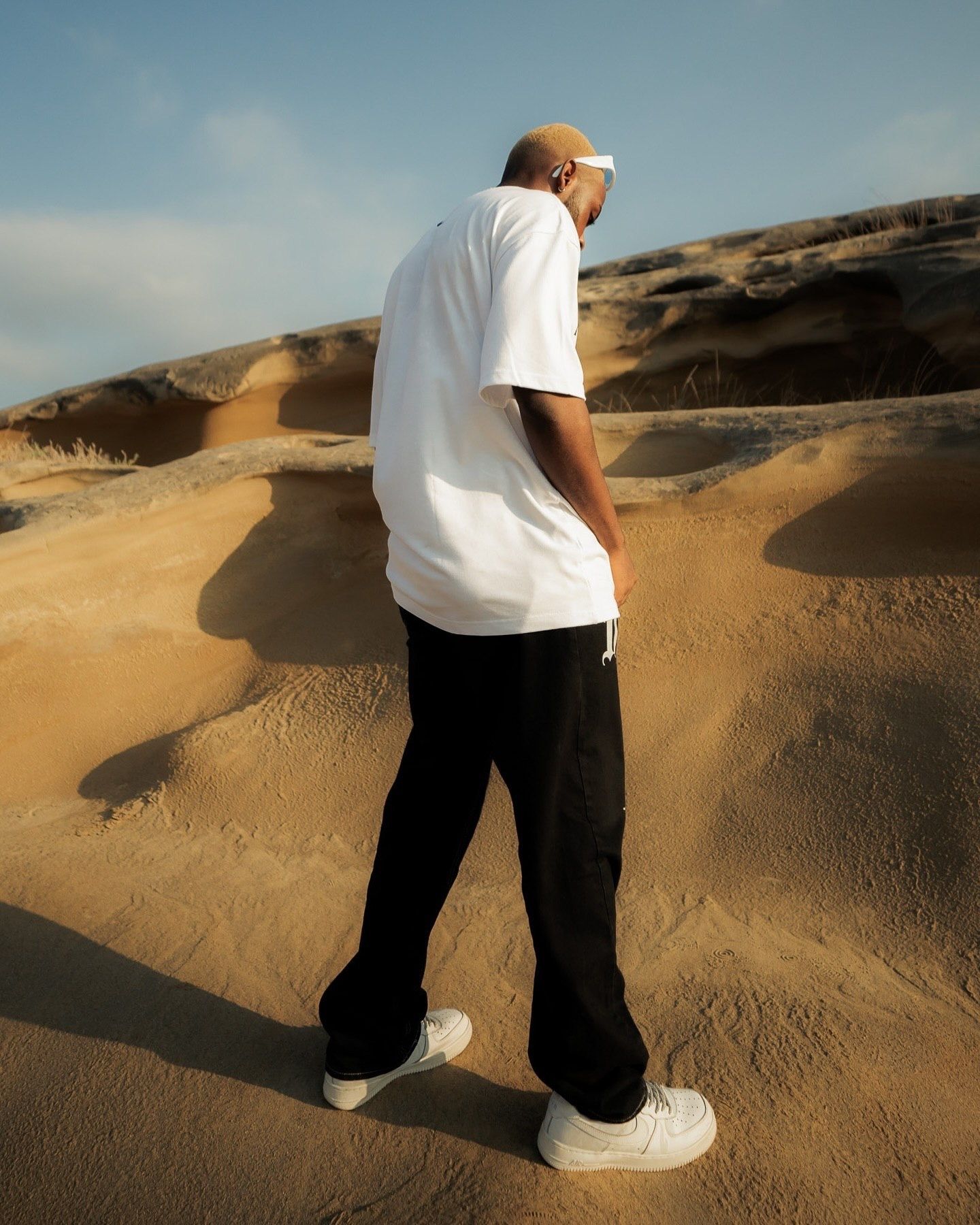 Man in white t-shirt, black pants, and white sneakers standing on golden sand dunes under a blue sky.