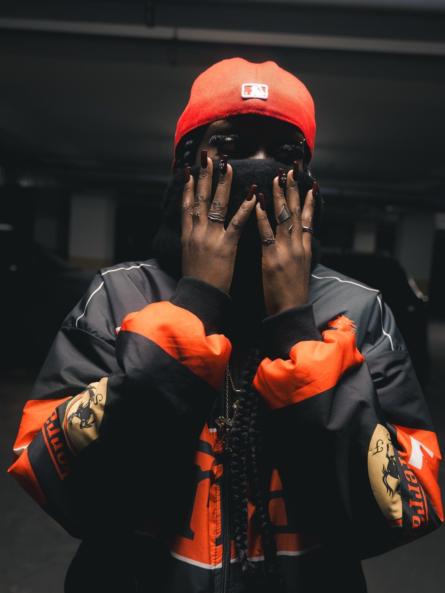 Person in red MLB beanie and orange bomber jacket covering face with ringed hands in dark parking garage.