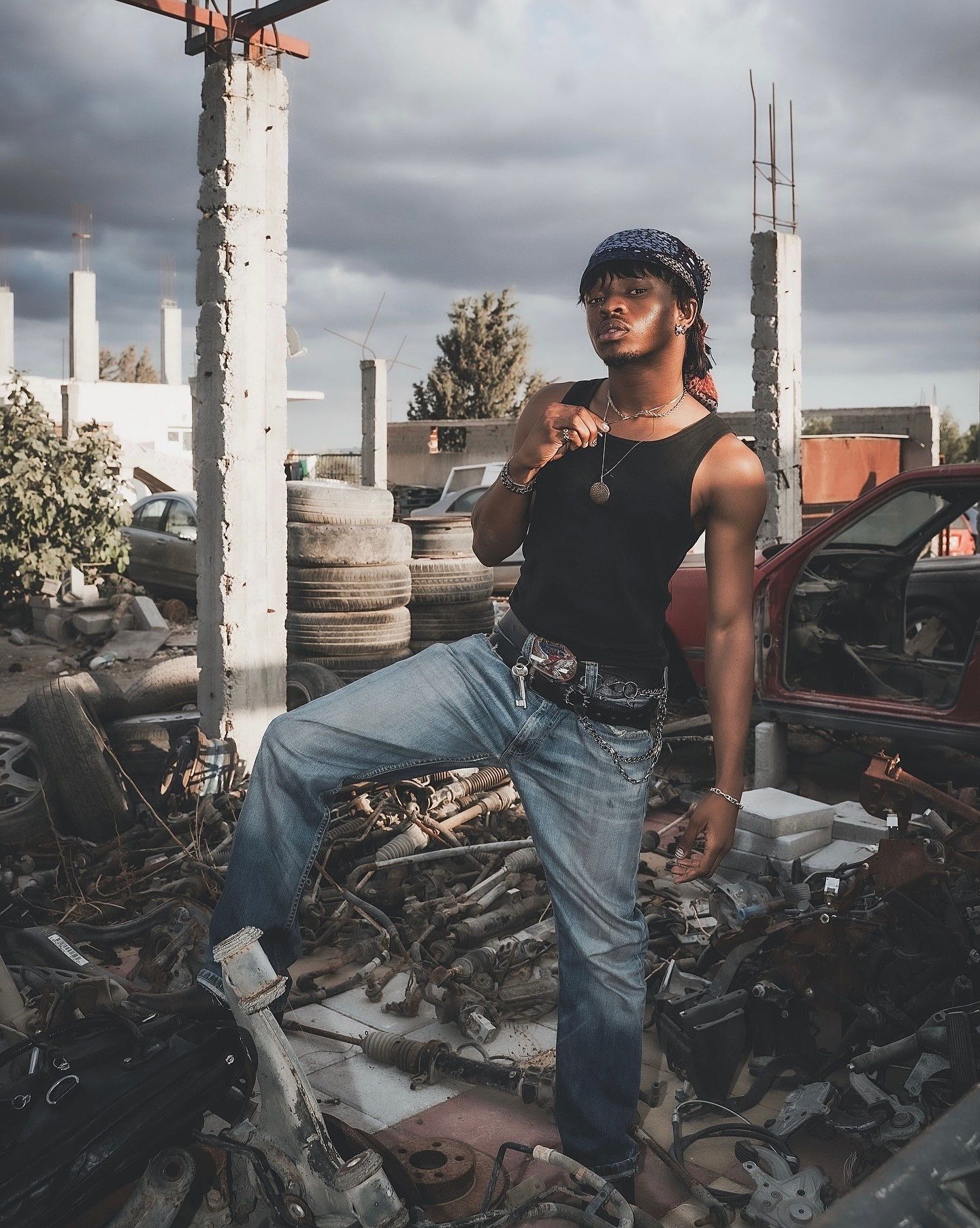 Young man in jeans and black tank top posing confidently in a junkyard under dramatic cloudy skies.