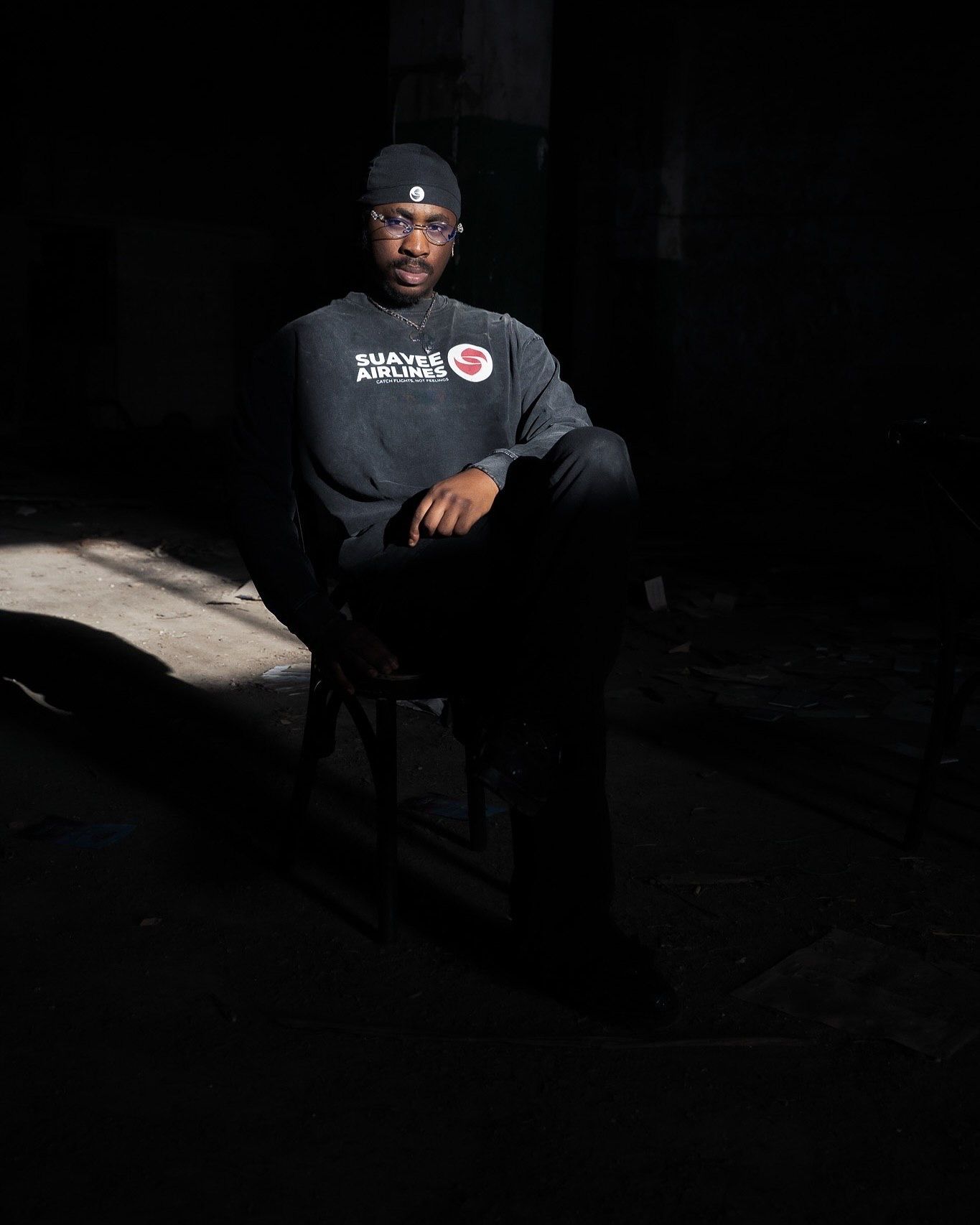 Man in dark beanie and Shayee Airlines sweatshirt seated on chair in dramatic low-key lighting portrait.