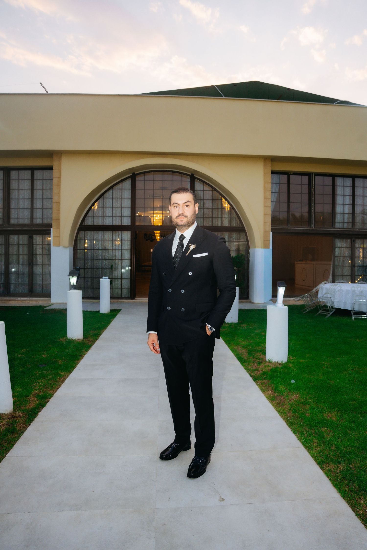 Man in black double-breasted suit standing on pathway outside elegant venue with arched windows at dusk.