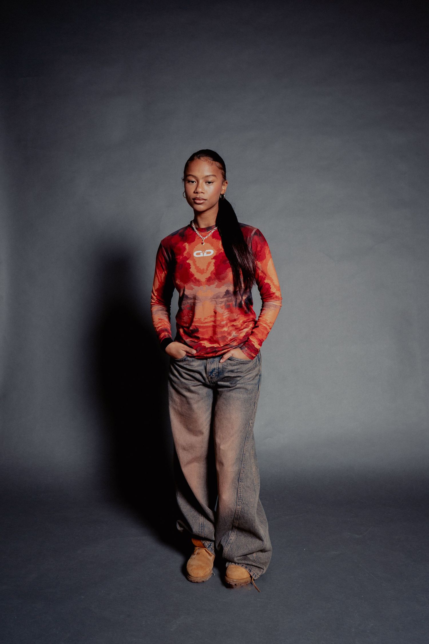 Young woman in red tie-dye long sleeve top, wide-leg gray jeans, and tan shoes standing against gray backdrop.