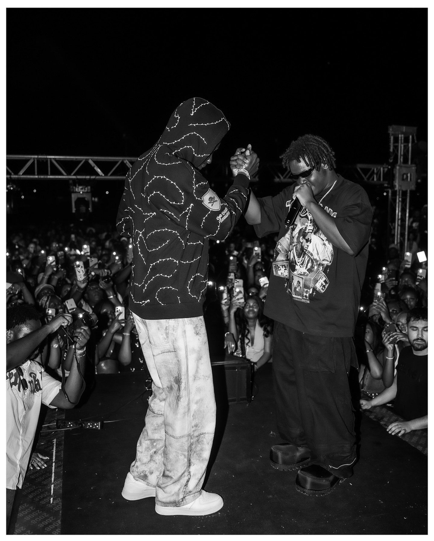 Two performers on stage at a packed night concert, one in a hoodie, both engaging the crowd in black and white.