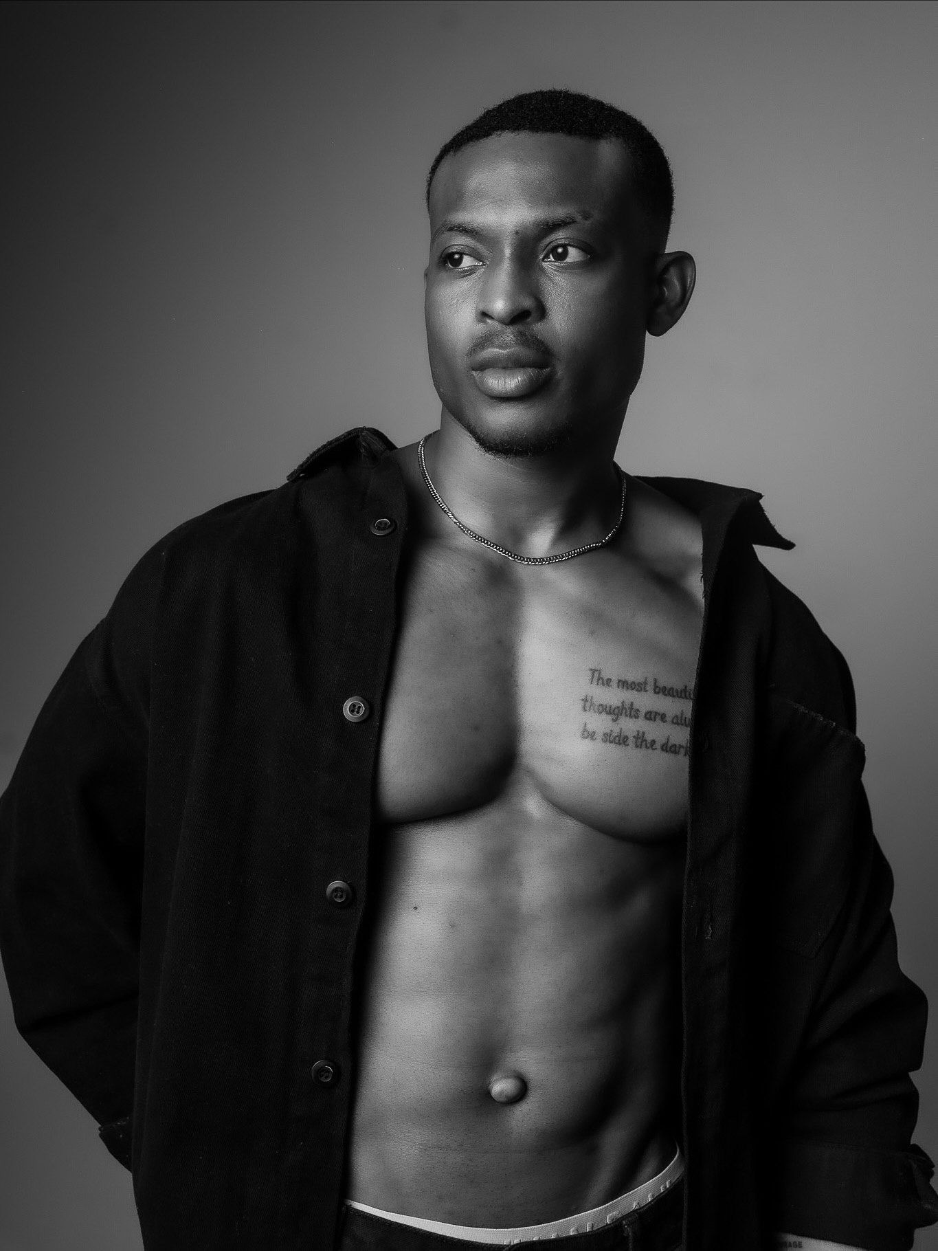 Black and white portrait of a young man in an open black jacket, showing chest tattoo, studio photography.