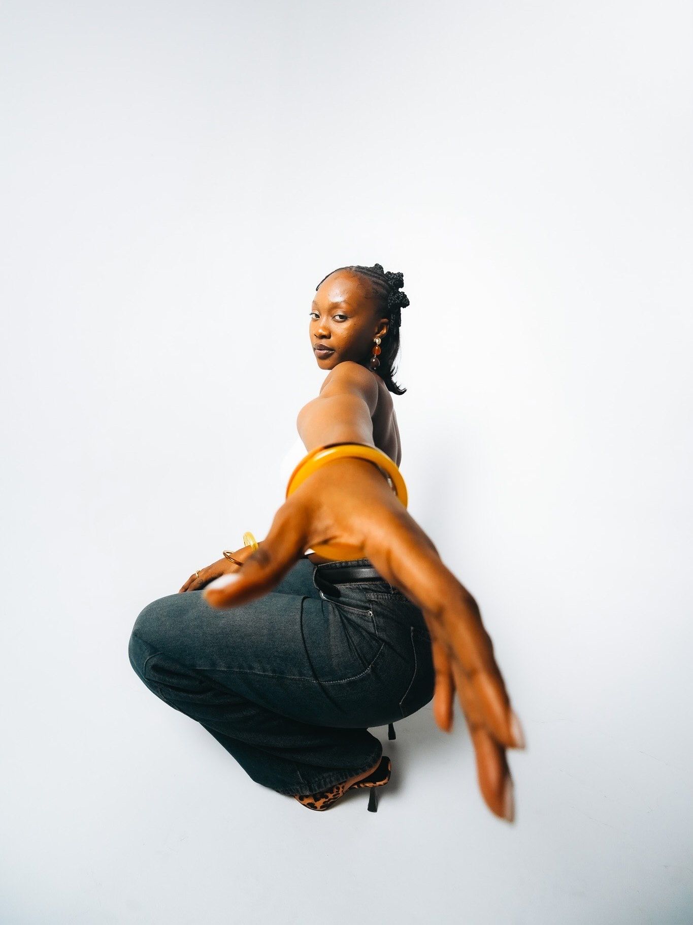 Black woman in yellow crop top and dark jeans posing in a crouched position against white backdrop.