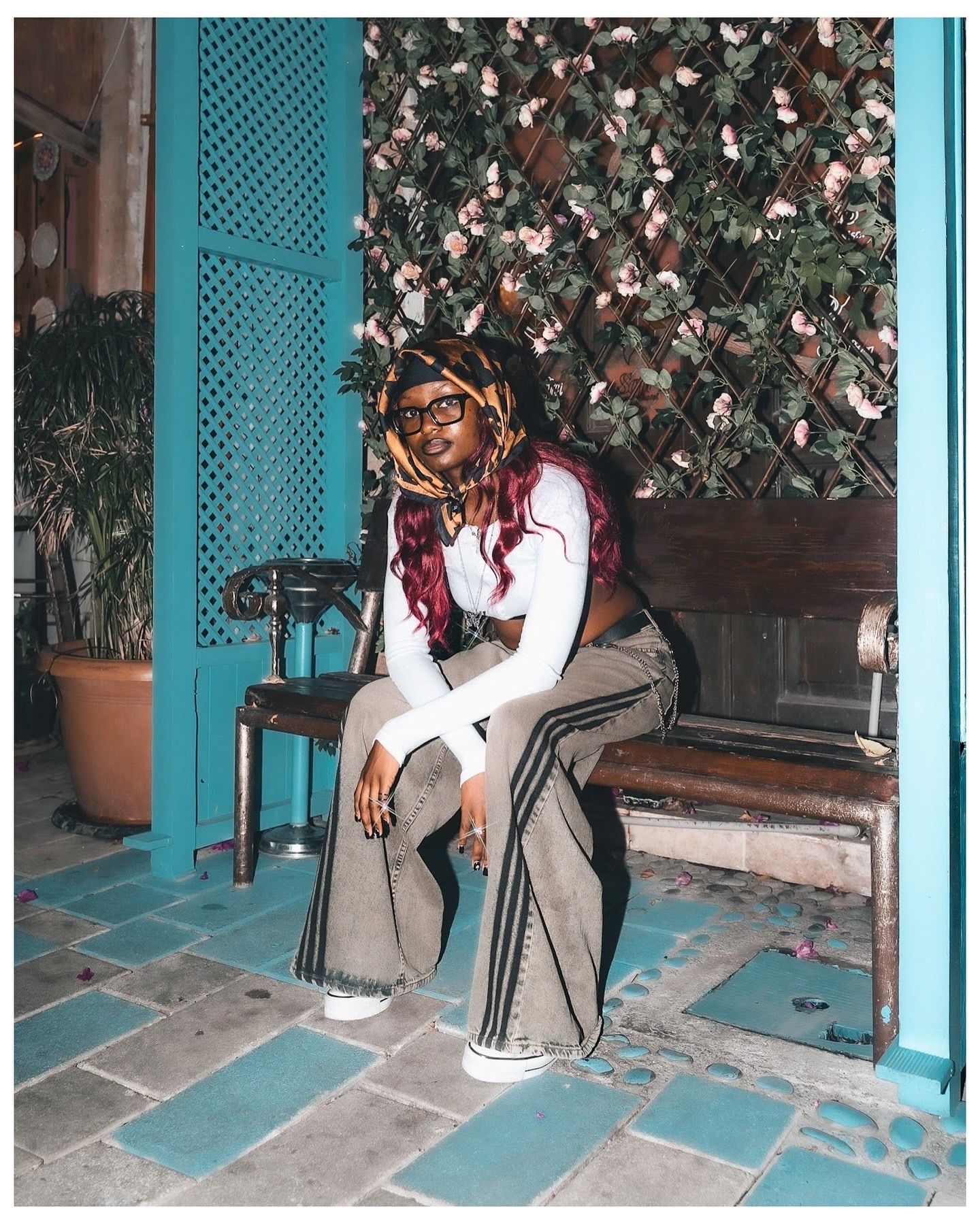 Young woman with colorful locs sits on a bench against a teal wall with floral greenery backdrop.